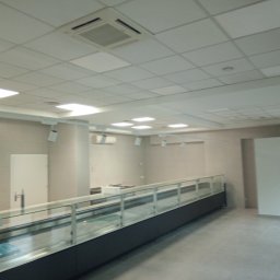 Tomasz Rogaczewski - Wnętrze sklepu lub lokalu usługowego w trakcie wykańczania: widoczna surowa posadzka, ściany w jasnych odcieniach, podwieszany sufit z oświetleniem, przeszklona lada chłodnicza oraz klimatyzacja.