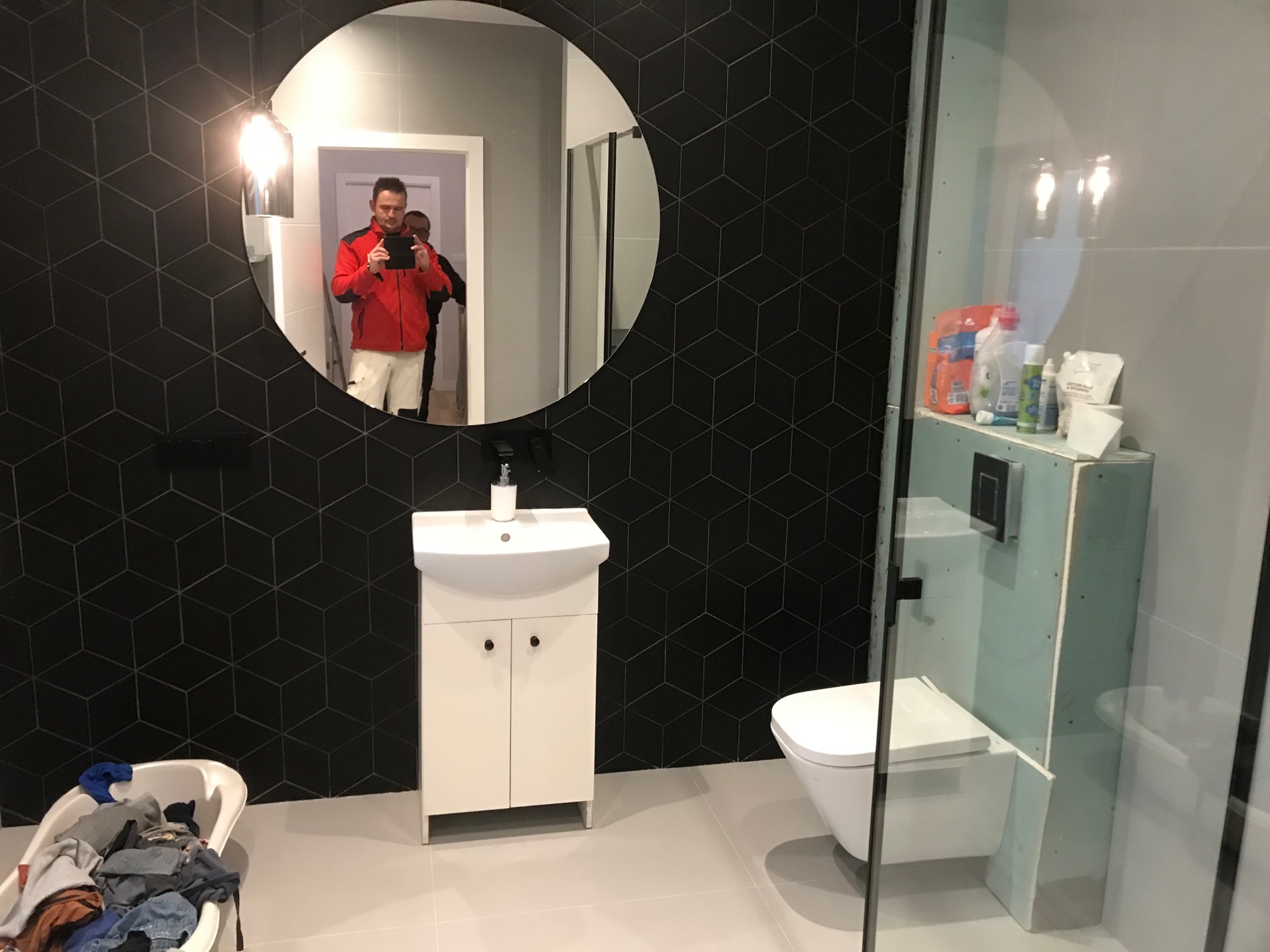 Nowoczesna łazienka z czarnymi płytkami w geometryczne wzory, okrągłe lustro odbijające osobę robiącą zdjęcie, biała umywalka z szafką i toaleta w trakcie instalacji za szklaną ścianką prysznicową.
