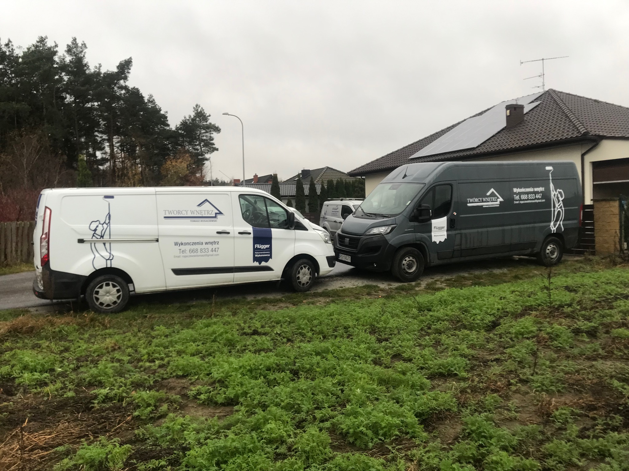 Dwa busy firmy wykończeniowej zaparkowane przed domem jednorodzinnym z panelami słonecznymi na dachu; na samochodach widnieje logo firmy i numer telefonu.