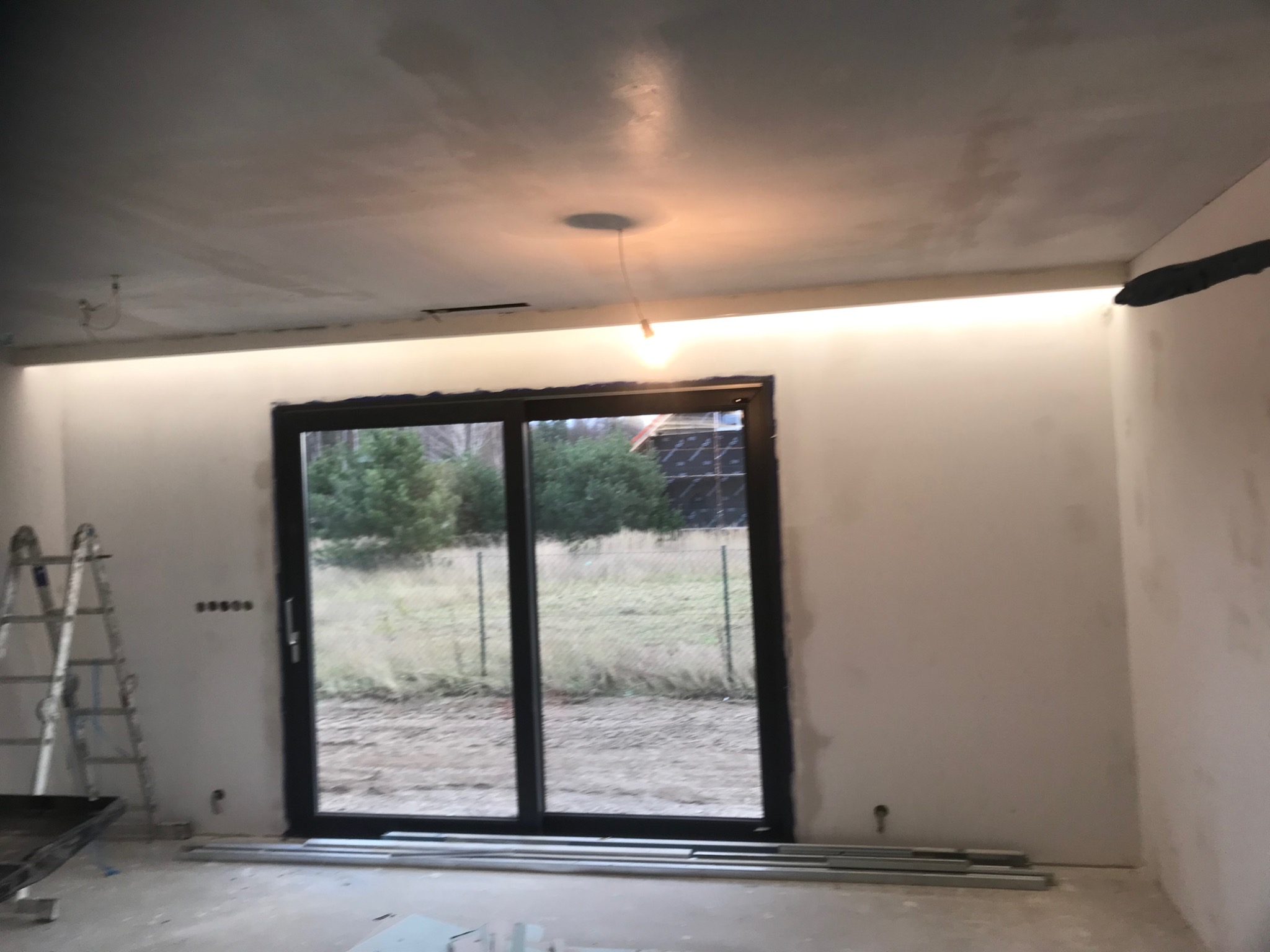 Surowy stan wnętrza z widocznymi śladami po szpachlowaniu ścian i sufitu, przygotowanego do dalszych prac wykończeniowych, z zamontowanymi przesuwnymi drzwiami balkonowymi i oświetleniem LED...