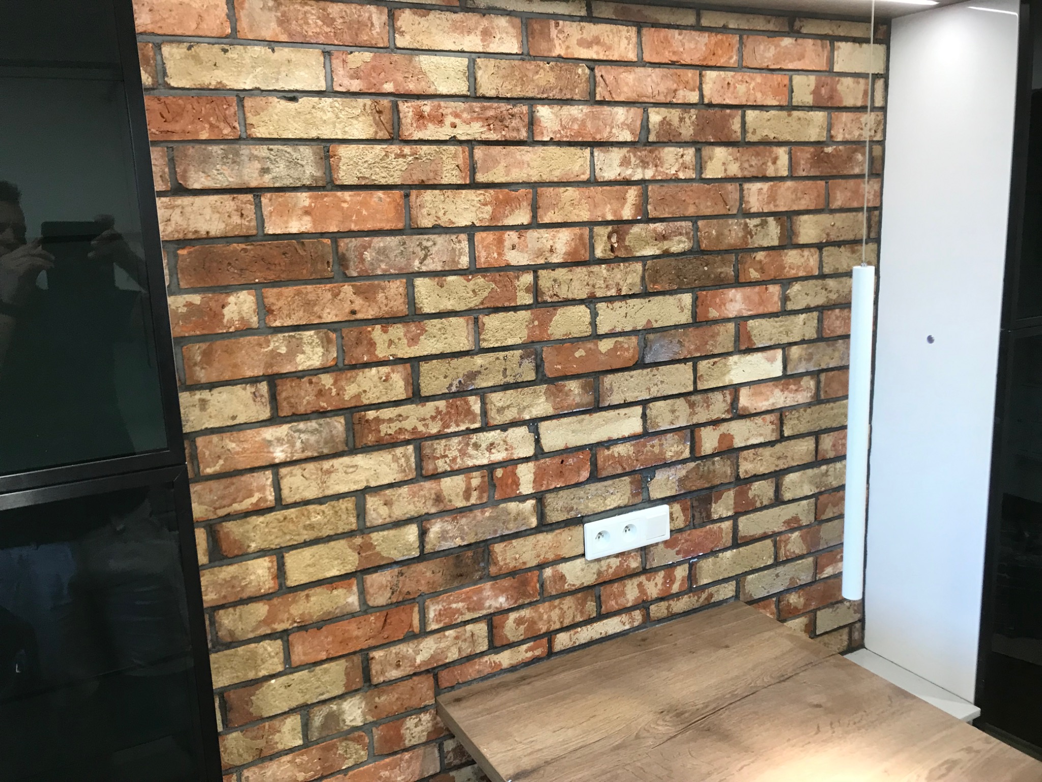 Ściana z cegły klinkierowej w odcieniach czerwieni i beżu, z widocznym gniazdkiem elektrycznym i fragmentem blatu drewnianego. W tle fragment czarnej ramy okiennej z odbiciem osoby robiącej zdjęcie.