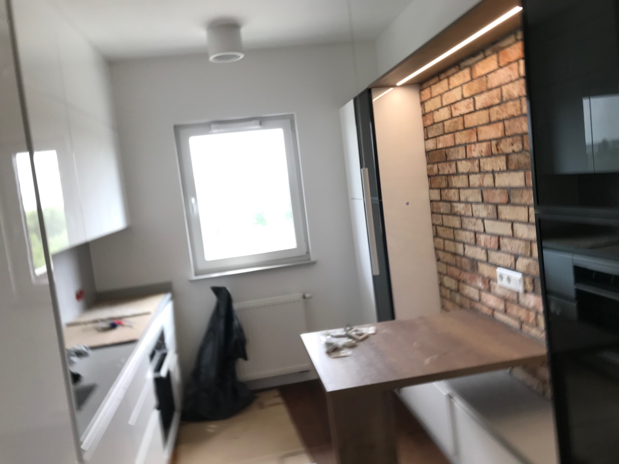 Wnętrze kuchni w trakcie remontu: białe szafki kuchenne, ceglana ściana z oświetleniem LED, okno i nieporządek na podłodze.