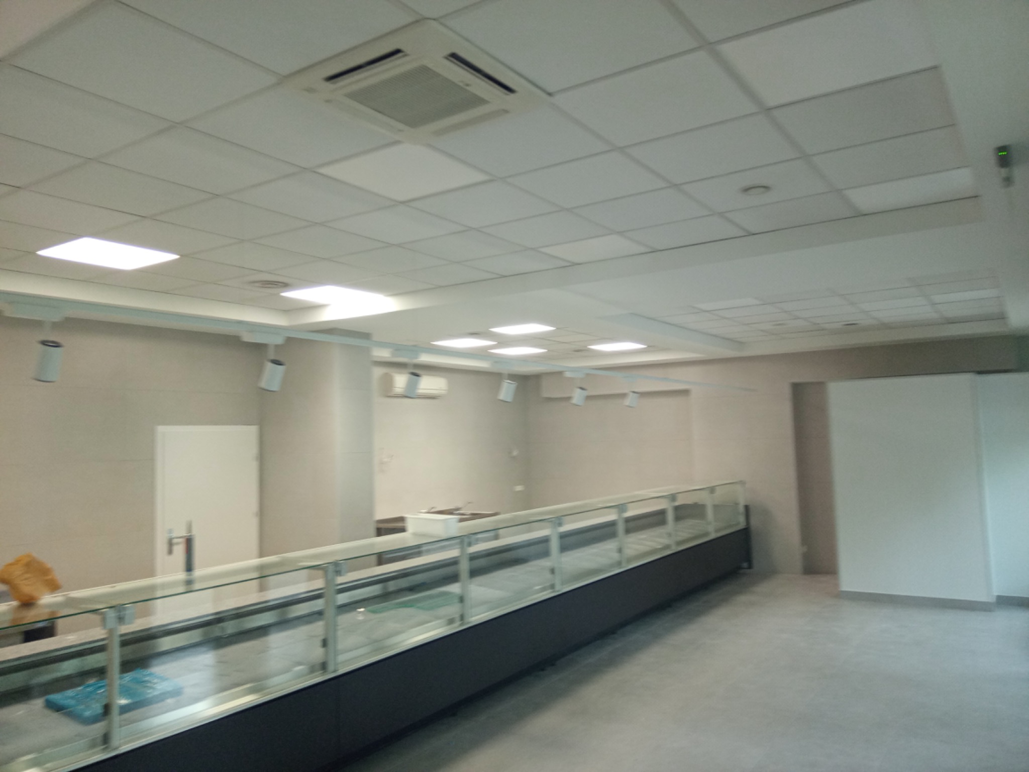 Wnętrze sklepu lub lokalu usługowego w trakcie wykańczania: widoczna surowa posadzka, ściany w jasnych odcieniach, podwieszany sufit z oświetleniem, przeszklona lada chłodnicza oraz klimatyzacja.