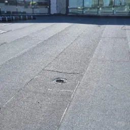 Szary dach pokryty papą termozgrzewalną z widocznym odpływem liniowym, butla gazowa i rolki papy w tle.