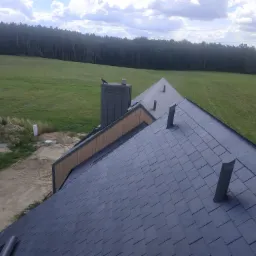 Widok z góry na nowy dach z ciemnych łupków, z kominem i wentylacją, na tle zielonego pola i lasu w pochmurny dzień.