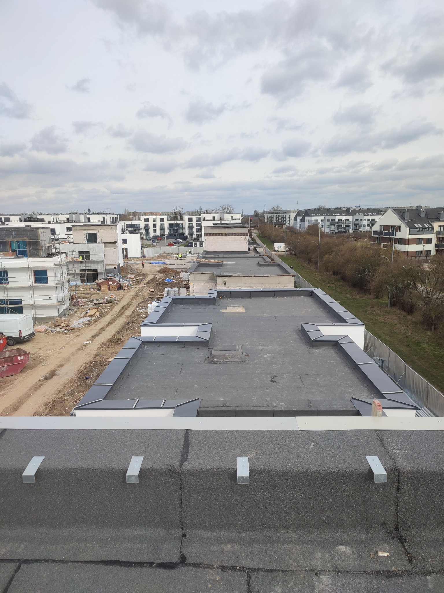 Widok z góry na płaski dach pokryty papą, z widocznymi obróbkami blacharskimi w kolorze szarym, w tle budynki w trakcie budowy i pochmurne niebo.