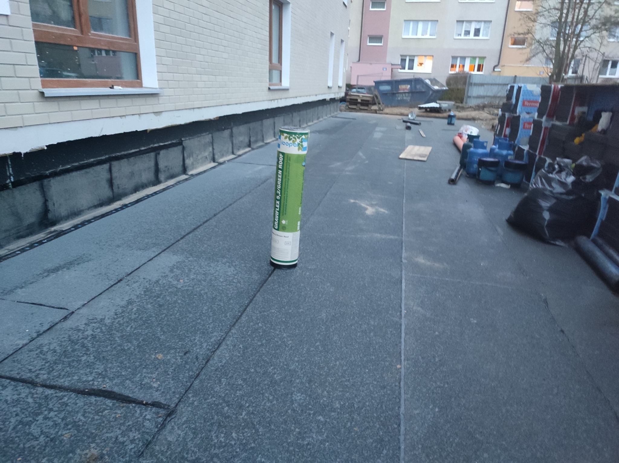 Wykończony dach płaski budynku z widocznym materiałem izolacyjnym na ścianie, rolka papy na dachu i narzędzia w tle.