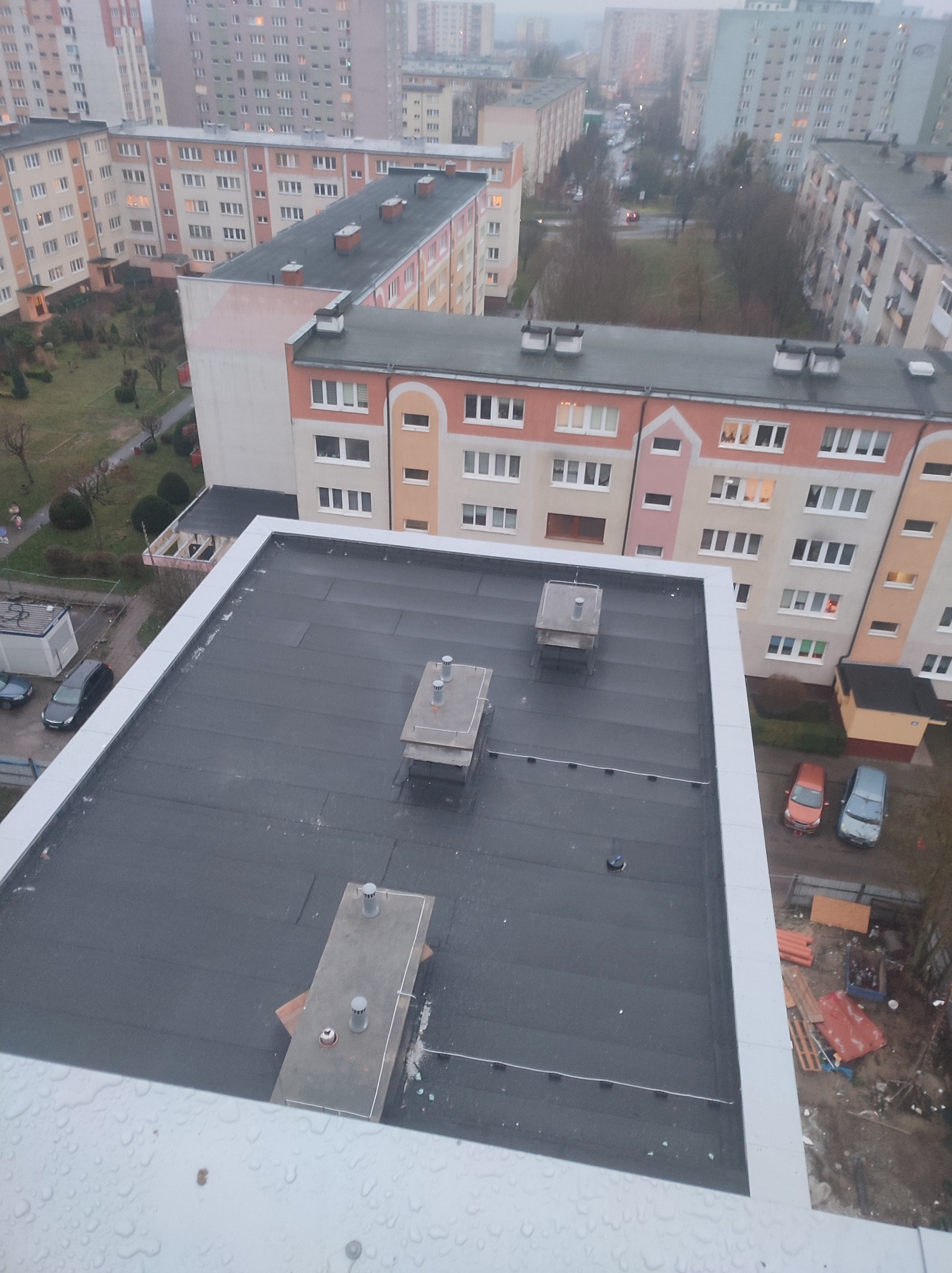 Widok z góry na płaski dach pokryty czarną papą termozgrzewalną, z trzema kominkami wentylacyjnymi; w tle bloki mieszkalne w szarej, pochmurnej pogodzie.