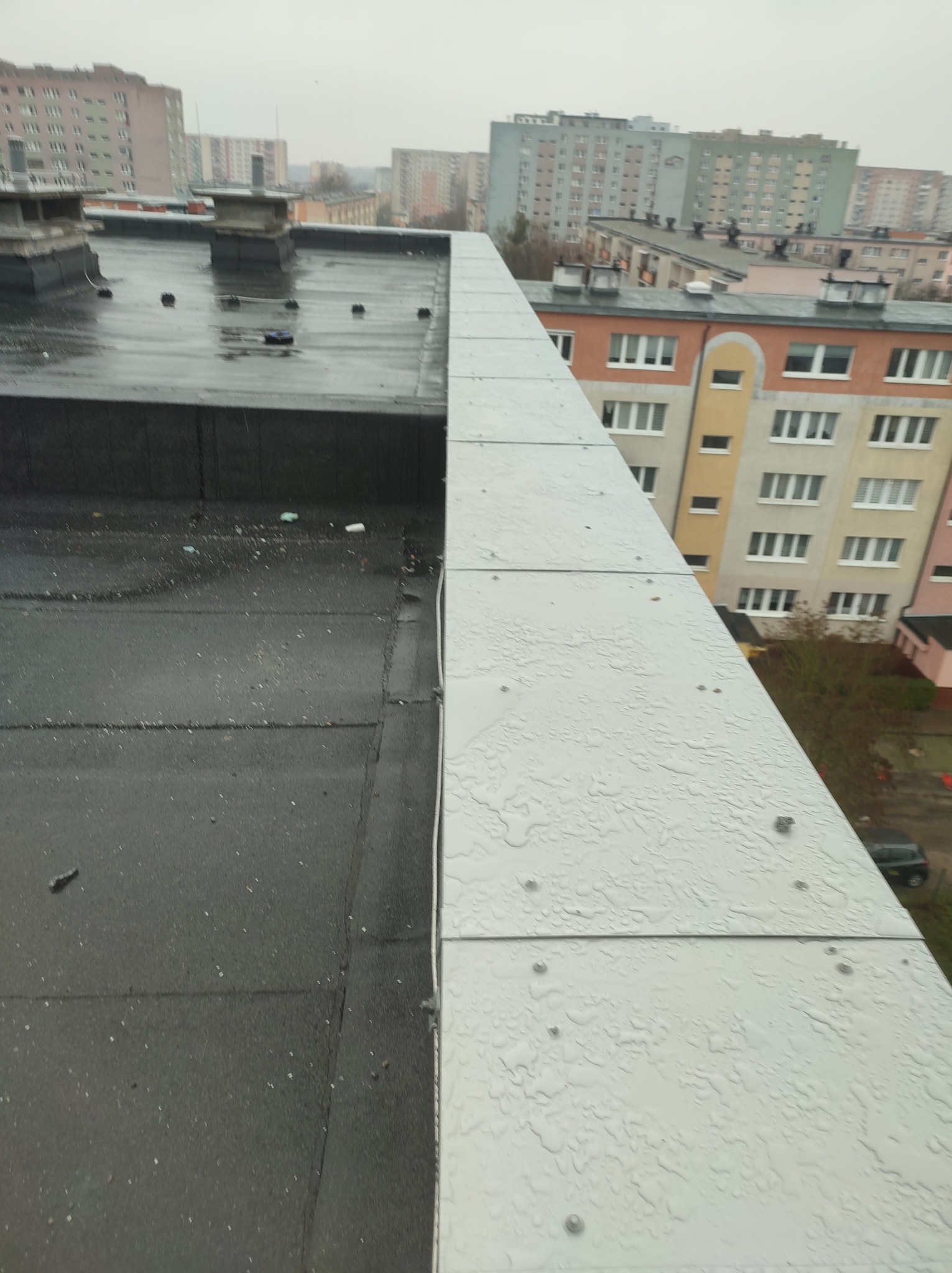 Widok na płaski, mokry dach z widoczną obróbką blacharską w kolorze srebrnym, na tle bloków mieszkalnych w pochmurny dzień.