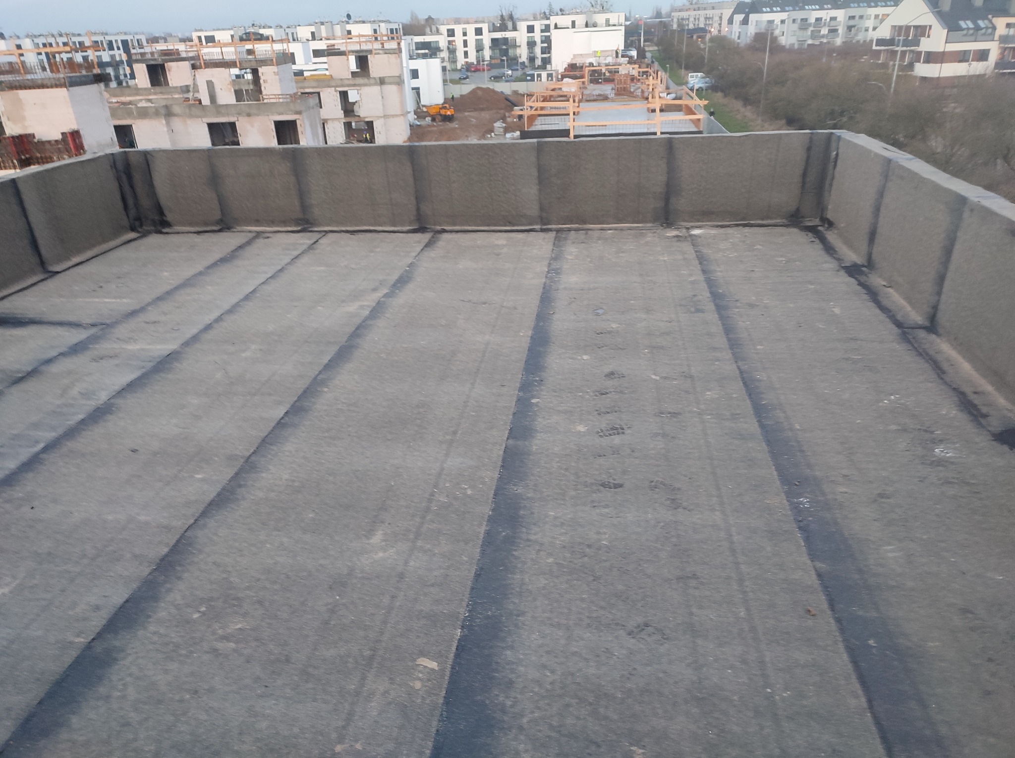 Płaski dach pokryty papą termozgrzewalną, z widoczną izolacją pionową attyki, na tle osiedla w budowie.