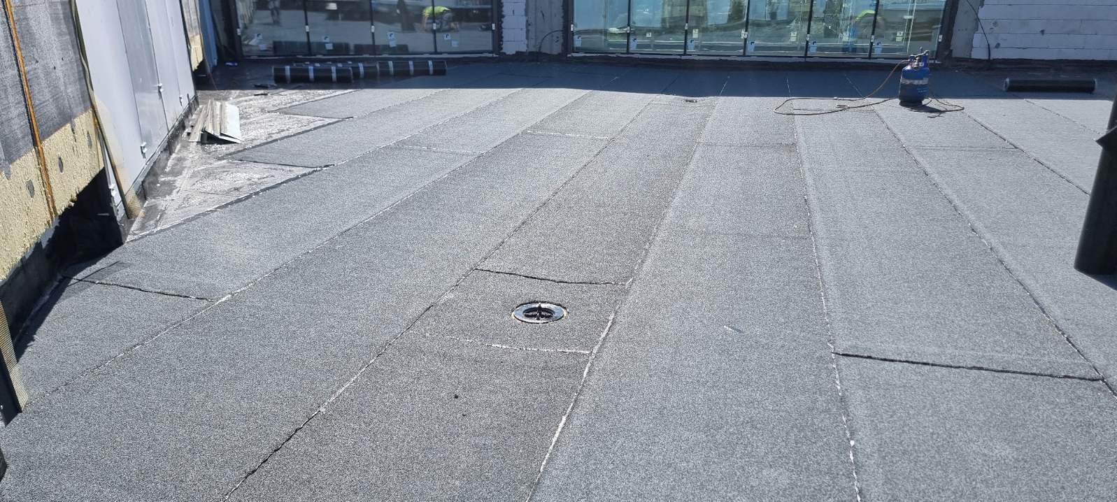 Szary dach pokryty papą termozgrzewalną z widocznym odpływem liniowym, butla gazowa i rolki papy w tle.