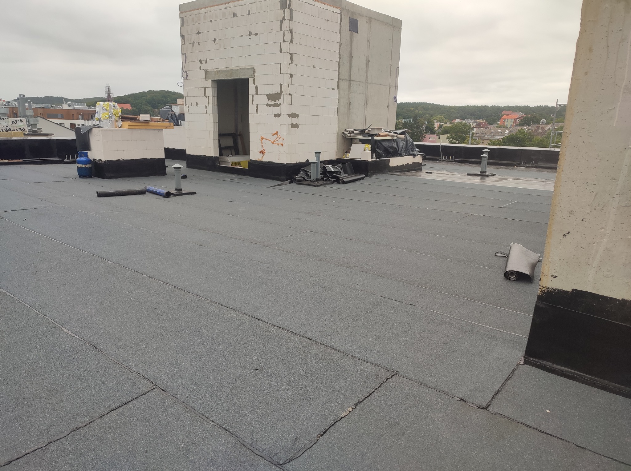 Szare, płaskie pokrycie dachowe z papy termozgrzewalnej na dachu budynku, widoczne kominy wentylacyjne, butla gazowa i fragmenty muru z pustaków.