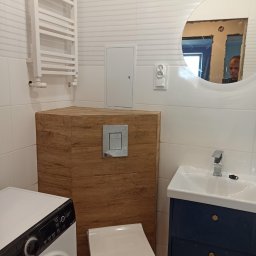 R-BUD - Nowoczesna łazienka z białymi płytkami imitującymi fale, podwieszaną toaletą z obudową z paneli drewnopodobnych, umywalką z szafką w kolorze granatowym i okrągłym lustrem, w którym odbija się osoba.