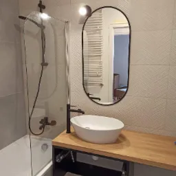 Nowoczesna łazienka z owalnym lustrem, białą umywalką nablatową na drewnianym blacie i czarną armaturą, wykończona płytkami w subtelny wzór jodełki.