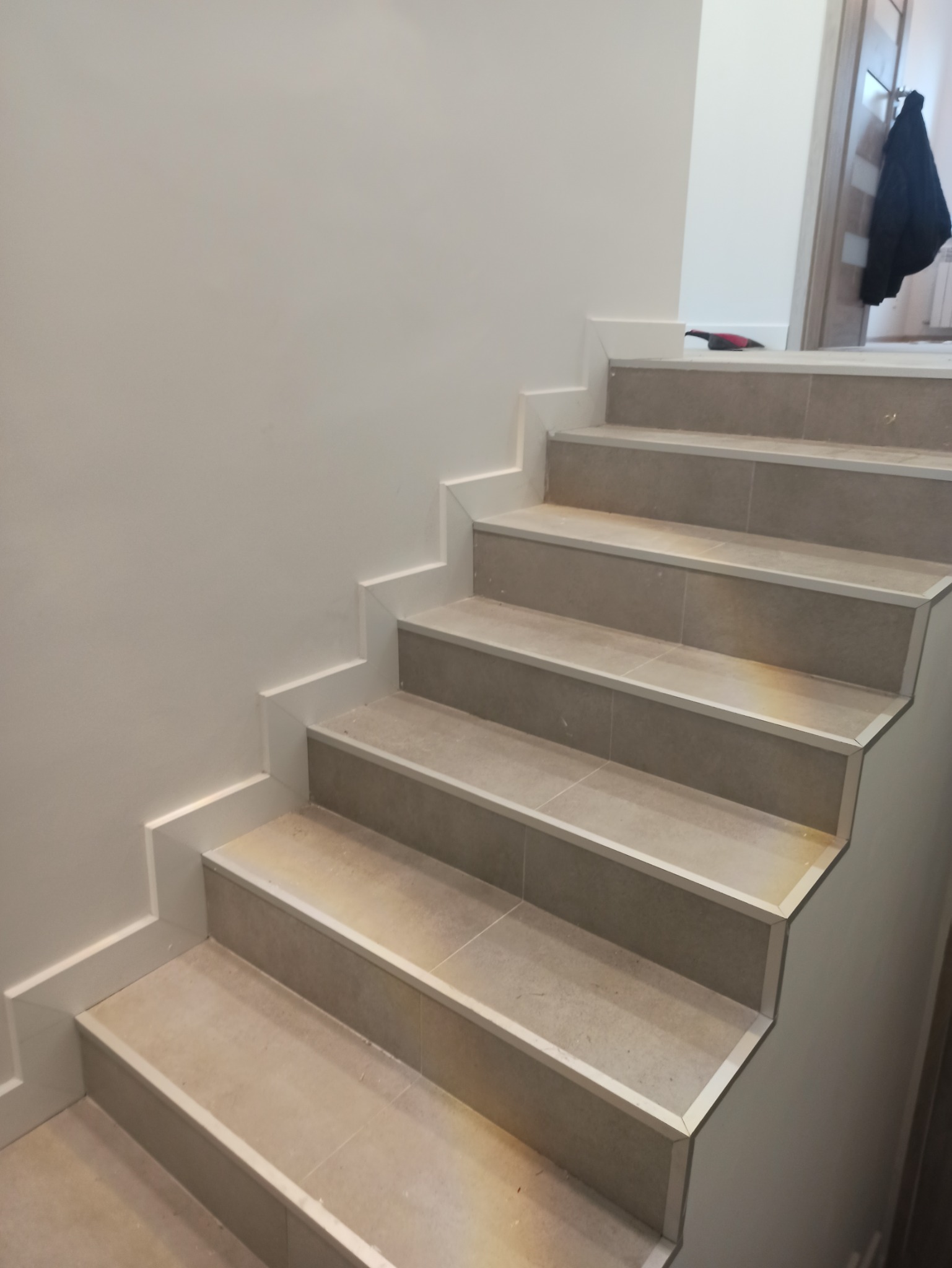 Schody wyłożone płytkami ceramicznymi w odcieniach beżu, z białymi listwami przypodłogowymi, widok z dołu, na górze schodów widoczny fragment czerwonego narzędzia, w tle drzwi i kurtka.