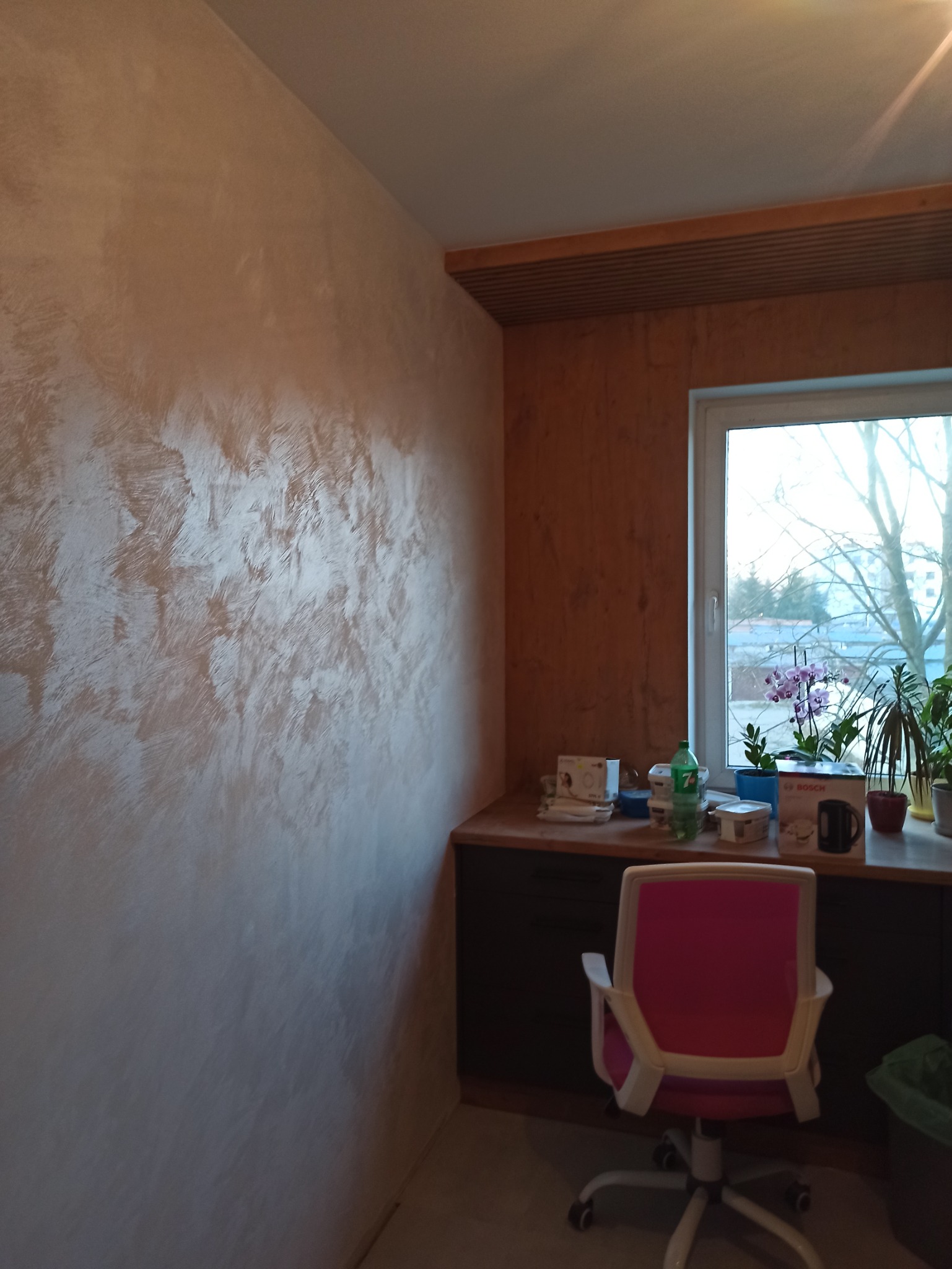 Ściana z dekoracyjną fakturą w odcieniach beżu i brązu, widoczna z perspektywy pokoju z biurkiem, oknem i roślinami doniczkowymi, oraz krzesłem biurowym z różowym oparciem.
