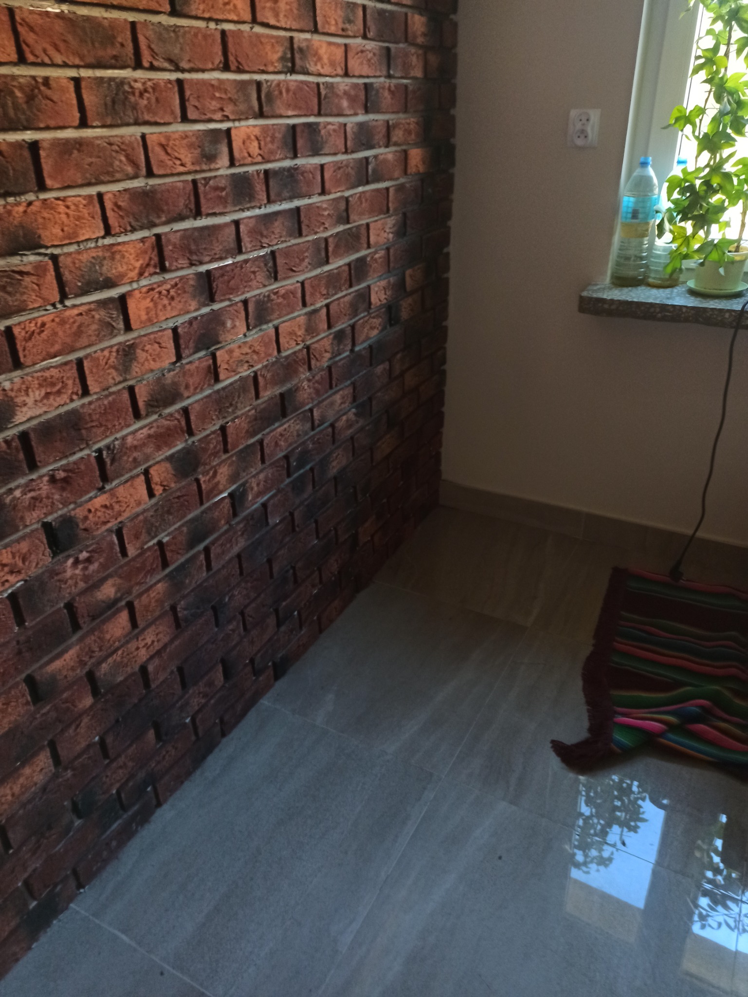 Ściana wyłożona cegłą klinkierową w rogu pokoju, szara podłoga z płytek i fragment okna z rośliną doniczkową.