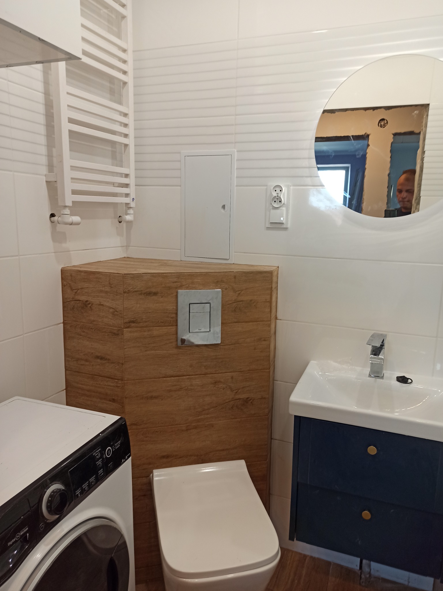 Nowoczesna łazienka z białymi płytkami imitującymi fale, podwieszaną toaletą z obudową z paneli drewnopodobnych, umywalką z szafką w kolorze granatowym i okrągłym lustrem, w którym odbija się osoba.