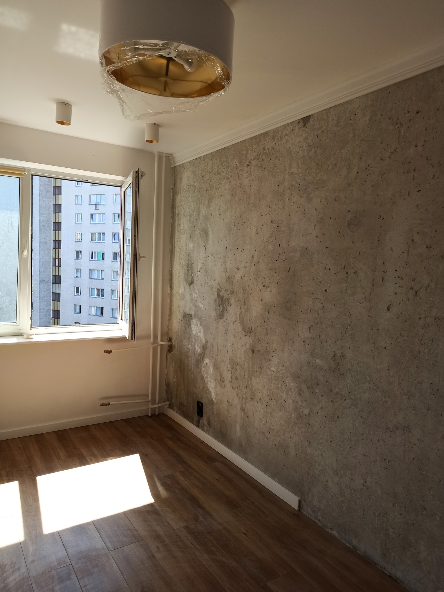 Pokój po remoncie: ściana z efektem betonu, drewnopodobna podłoga, białe listwy przypodłogowe, okno z widokiem na blok, lampa sufitowa w folii ochronnej.