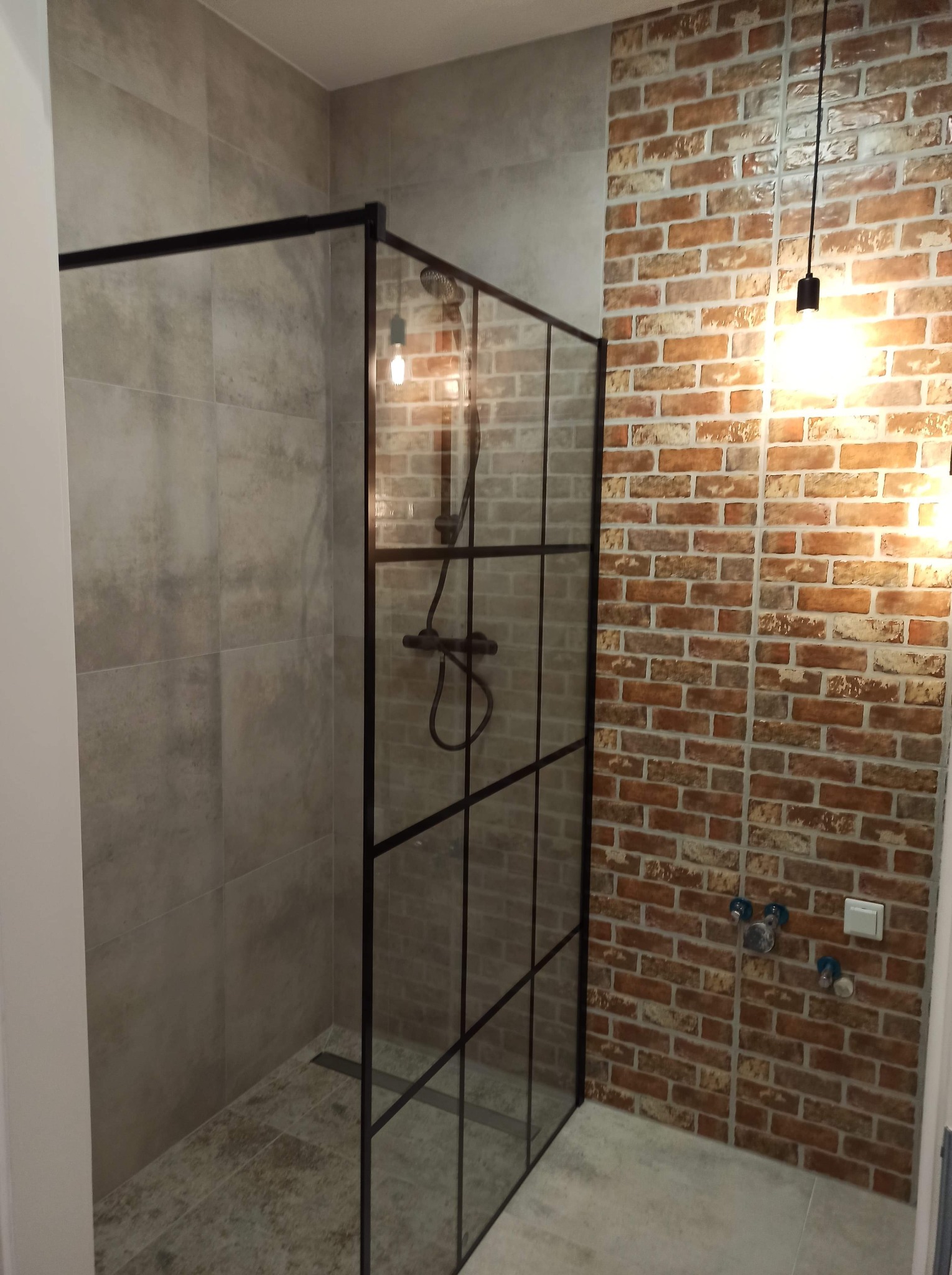 Nowoczesna kabina prysznicowa z czarnymi szprosami na tle ściany z cegły i szarych płytek, oświetlona wiszącymi lampami z żarówkami Edison.