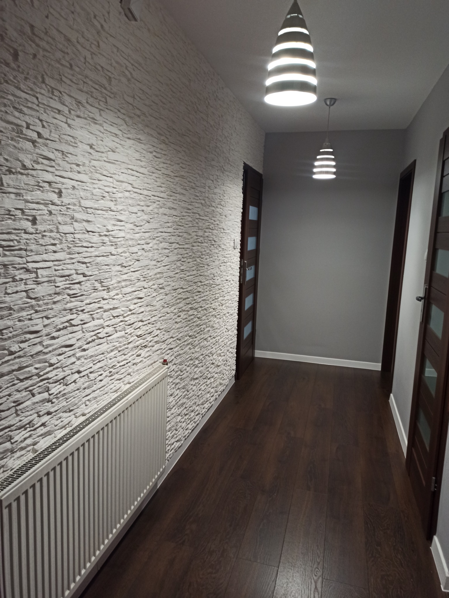 Korytarz z białymi płytkami imitującymi kamień na ścianie, ciemnymi panelami podłogowymi i dwoma nowoczesnymi lampami wiszącymi.