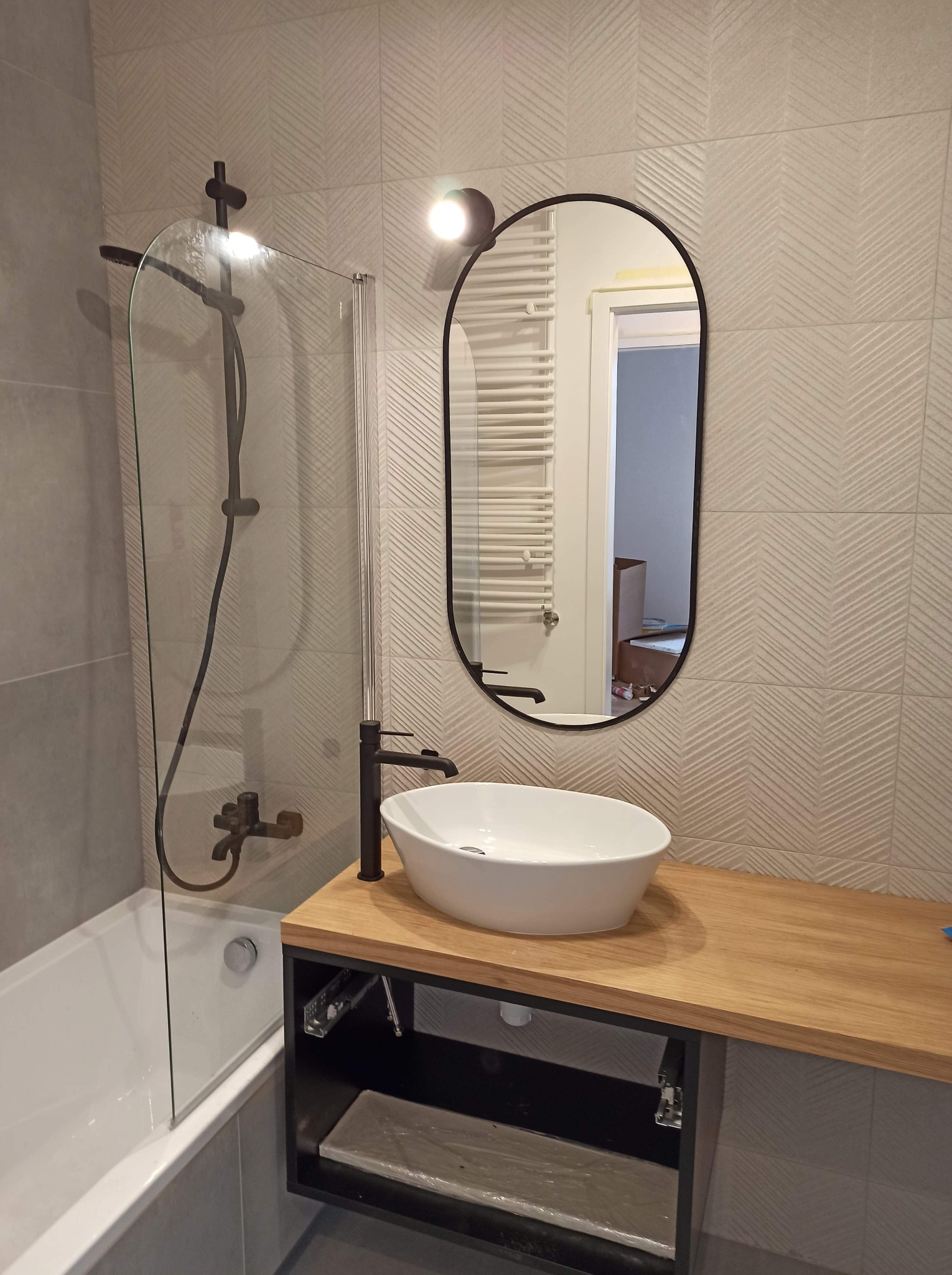 Nowoczesna łazienka z owalnym lustrem, białą umywalką nablatową na drewnianym blacie i czarną armaturą, wykończona płytkami w subtelny wzór jodełki.