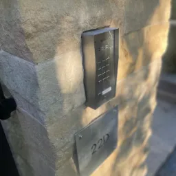 Zewnętrzny panel domofonowy z klawiaturą numeryczną i tabliczką z numerem domu 22D, zamontowany na kamiennym słupku ogrodzenia.