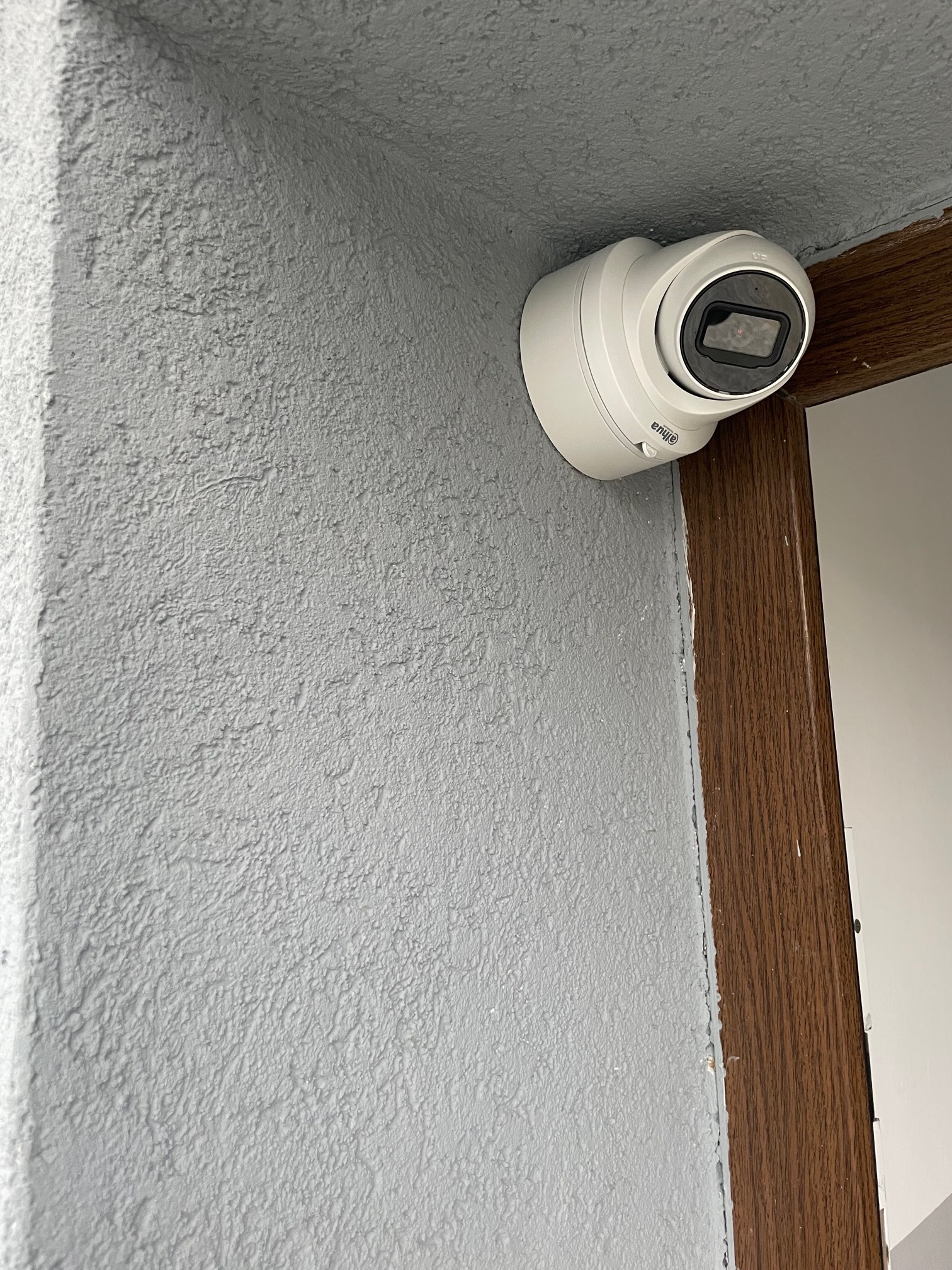 Kamera kopułkowa marki Dahua zamontowana na szarej, teksturowanej ścianie w rogu pomieszczenia, obok drewnianej framugi drzwi.