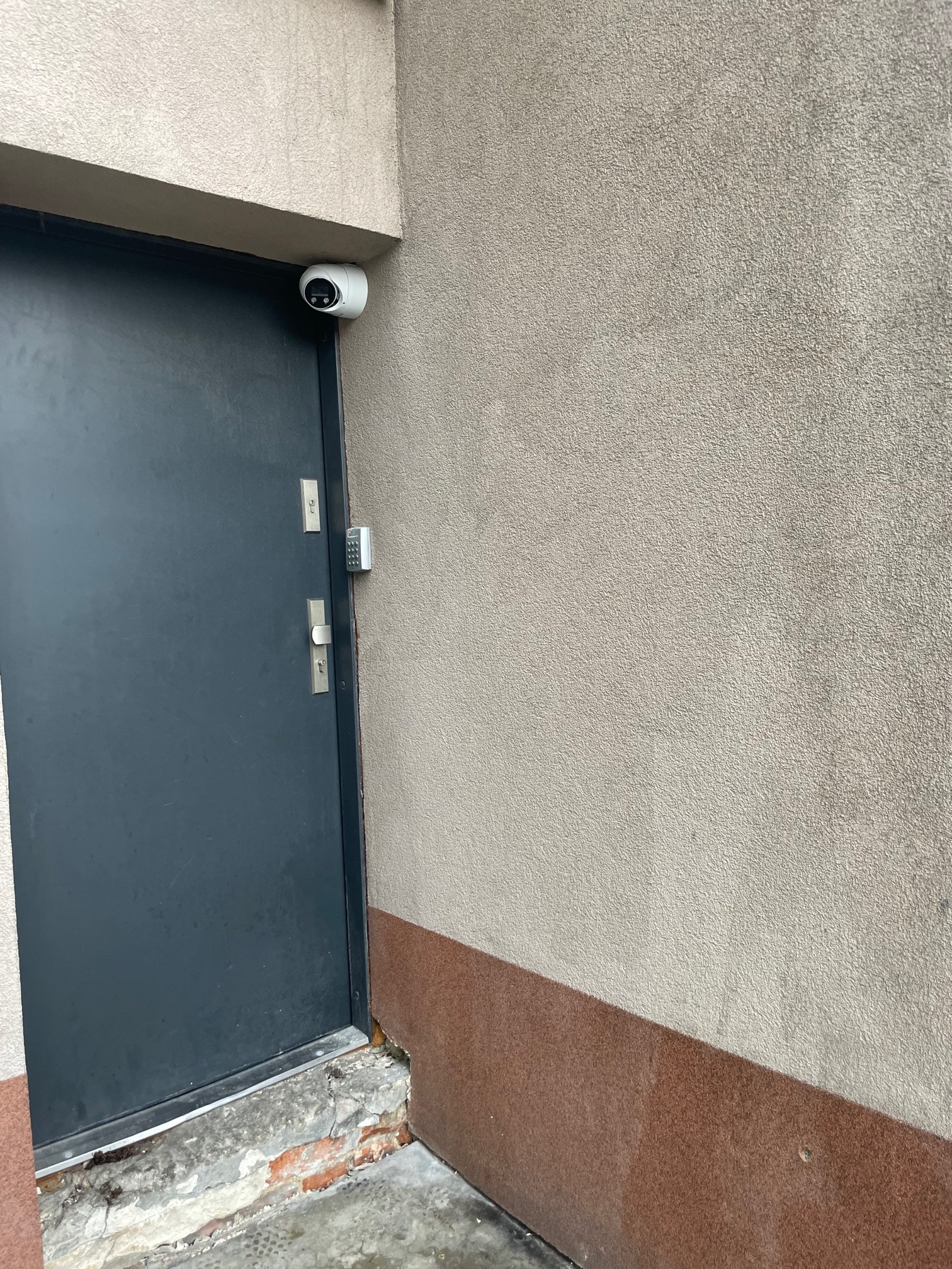 Zewnętrzna kamera monitoringu zamontowana nad szarą metalową bramą wejściową z panelem numerycznym, na tle beżowej ściany z brązowym cokołem.