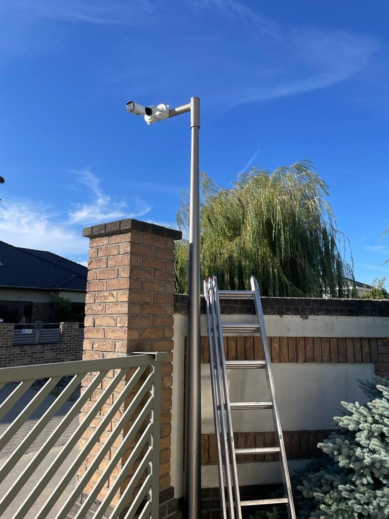 Dwie kamery CCTV zamontowane na metalowym słupie obok ceglanego słupka ogrodzeniowego, drabina oparta o słupek i słup, błękitne niebo w tle.