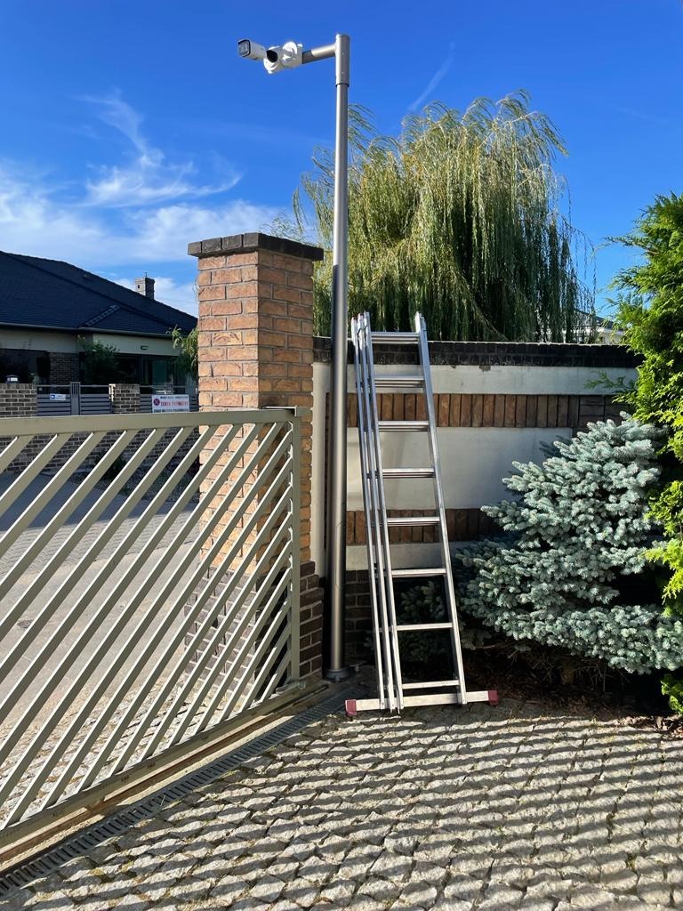 Dwie kamery monitoringu na wysokim słupie obok metalowej bramy wjazdowej, drabina oparta o słupek, widok z dołu w słoneczny dzień.