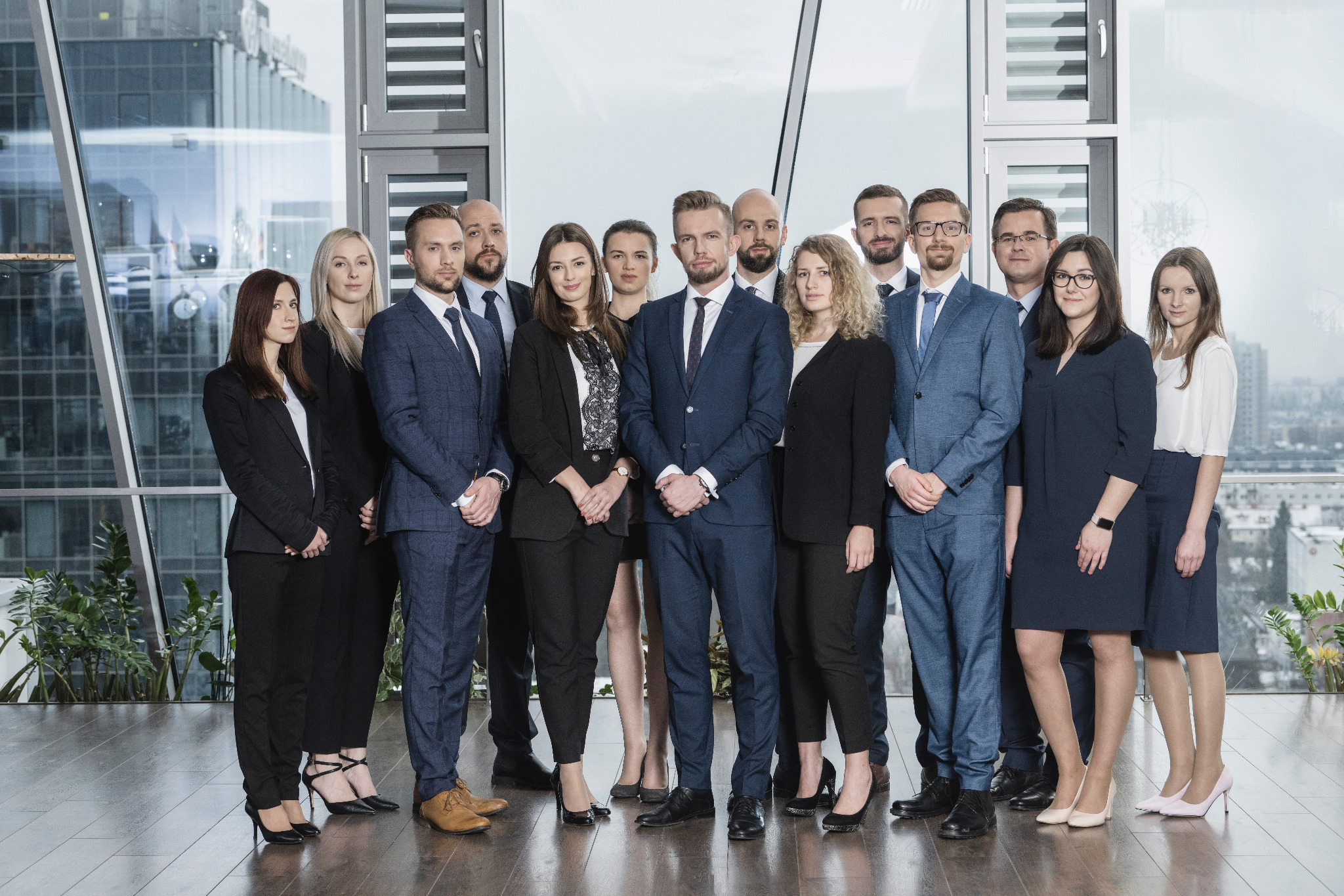 Zgrana grupa prawników w garniturach pozuje na tle okien wieżowca, widoczne detale ubioru i zróżnicowane fryzury.