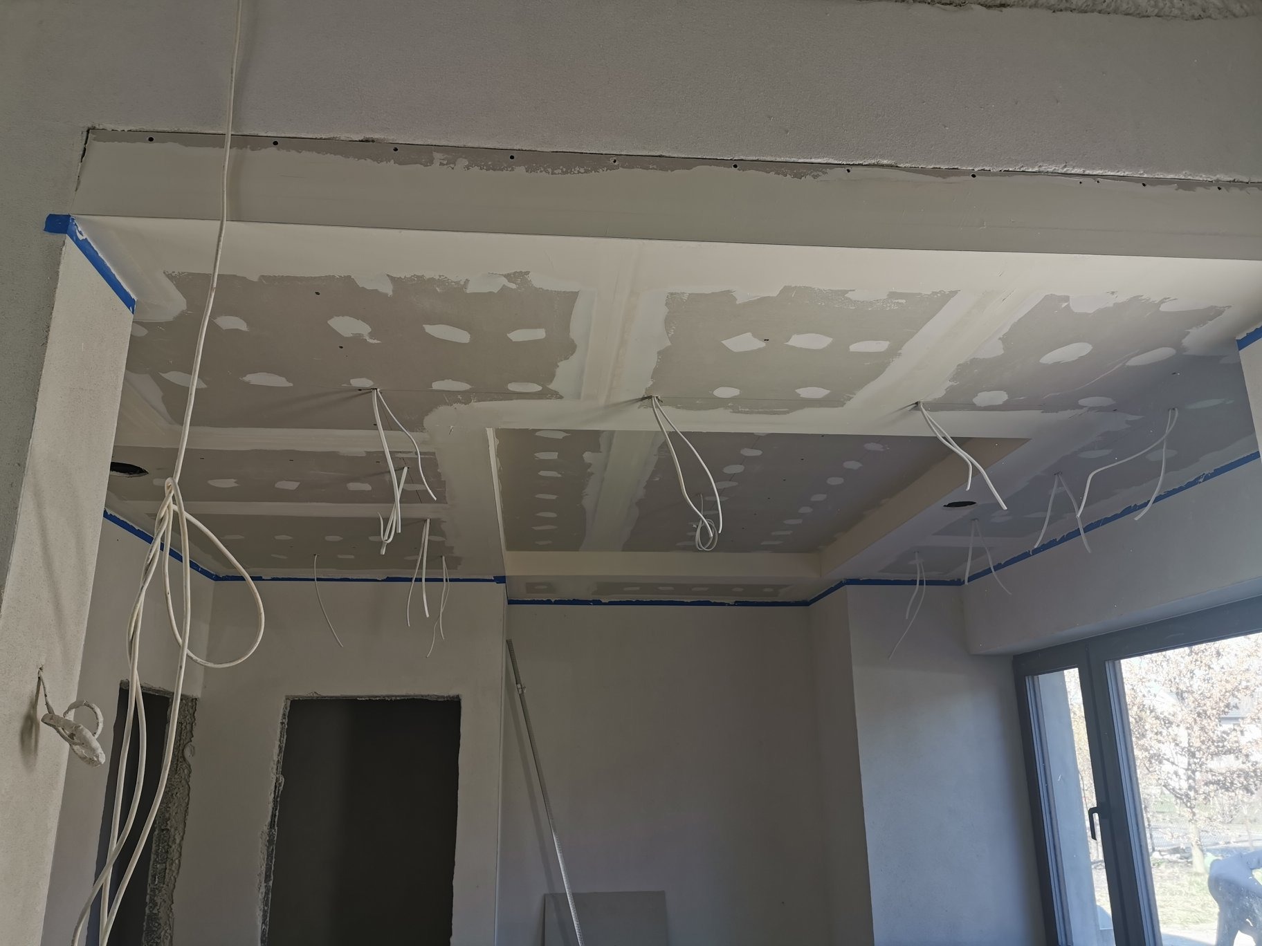 Wnętrze w trakcie prac wykończeniowych: sufit podwieszany z płyt gipsowo-kartonowych, widoczne kable elektryczne, ściany przygotowane do malowania, zabezpieczone taśmą malarską.