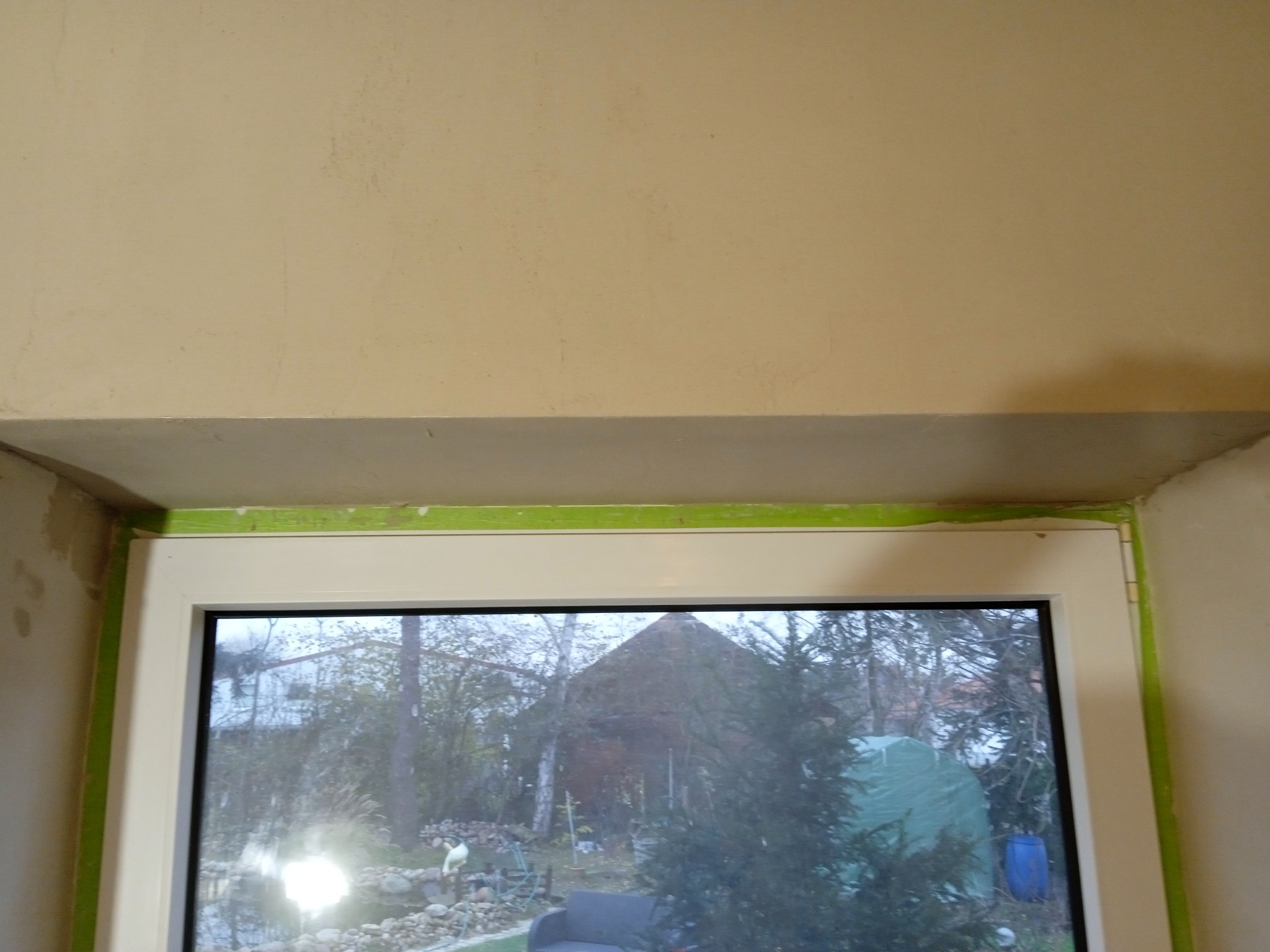 Przygotowanie ościeżnicy okiennej do malowania, zabezpieczona zieloną taśmą malarską, widoczne ślady szpachlowania na ścianie.