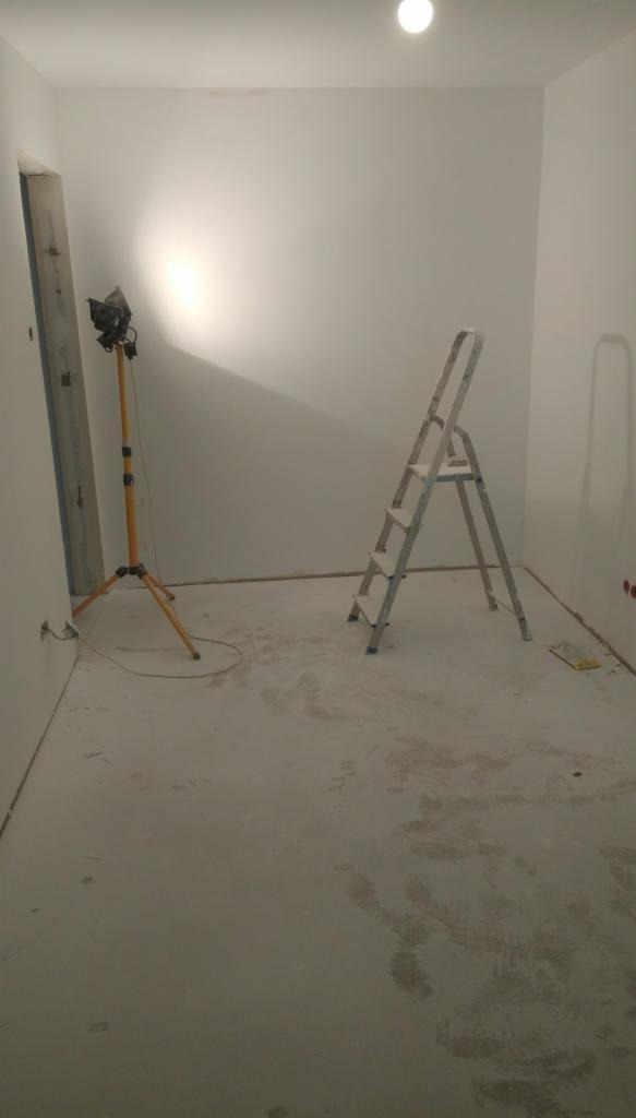 Pokój w trakcie prac wykończeniowych: ściany pomalowane na biało, widoczna drabina aluminiowa, lampa halogenowa na statywie i ślady farby na podłodze.