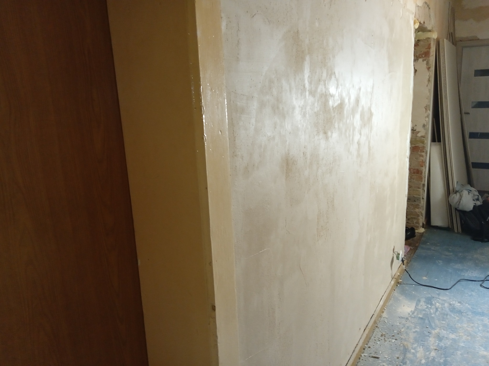 Świeżo nałożona, mokra warstwa szpachli na ścianie w trakcie prac wykończeniowych, widoczna fuga między ścianą a futryną drzwi, podłoga zabezpieczona folią.