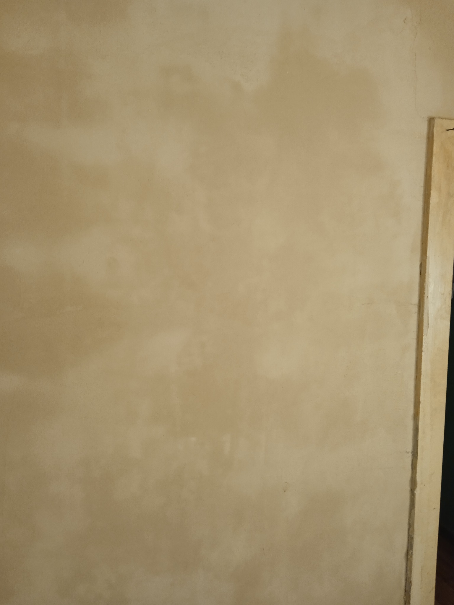 Ściana po szpachlowaniu, z widocznymi smugami i nierównościami, przygotowana do malowania, z fragmentem drewnianej ościeżnicy drzwiowej po prawej stronie.