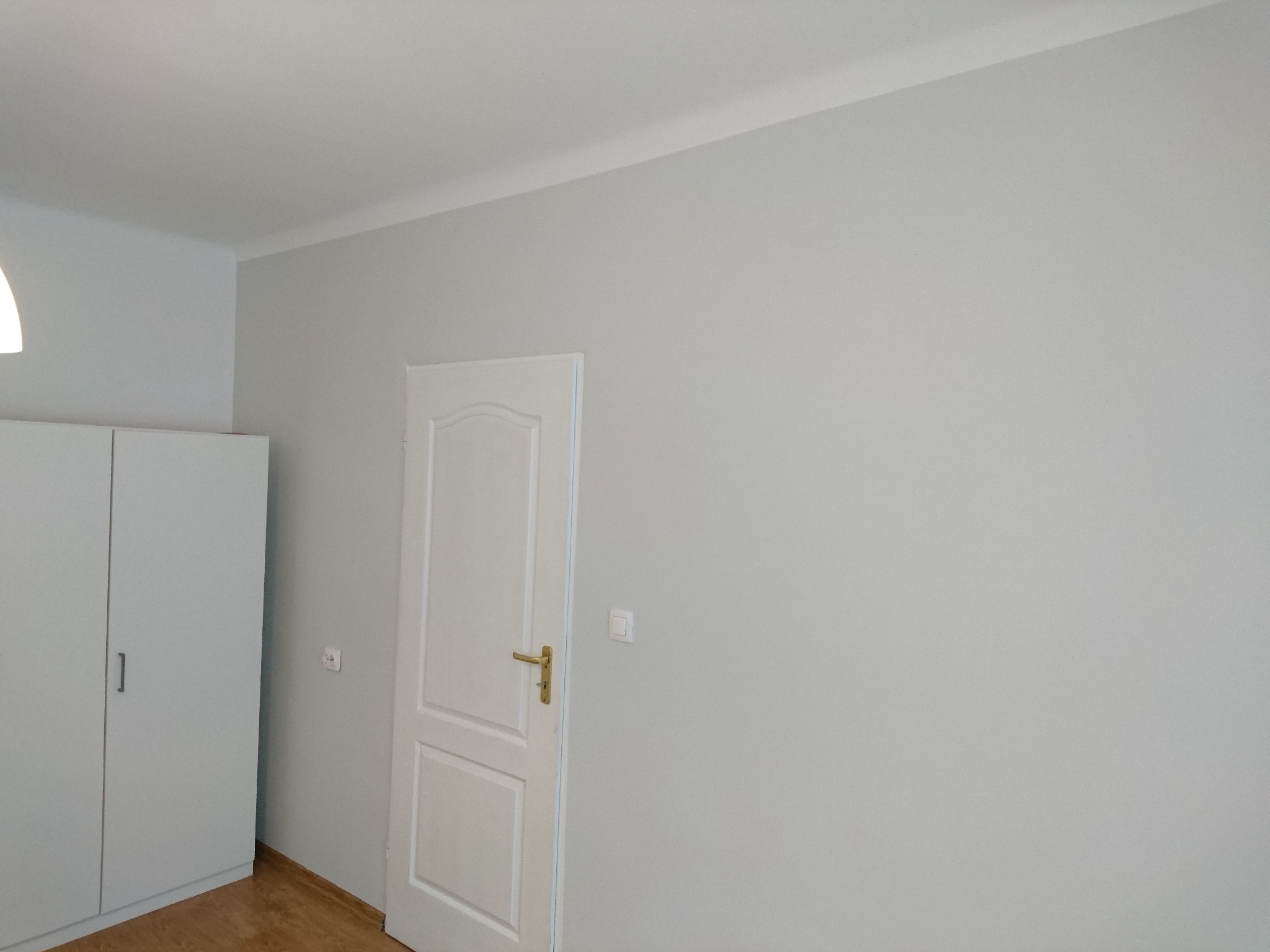 Jasnoszary pokój po odświeżeniu: białe drzwi z złotą klamką, biała szafa z minimalistycznym uchwytem, gładkie ściany i drewniana podłoga.