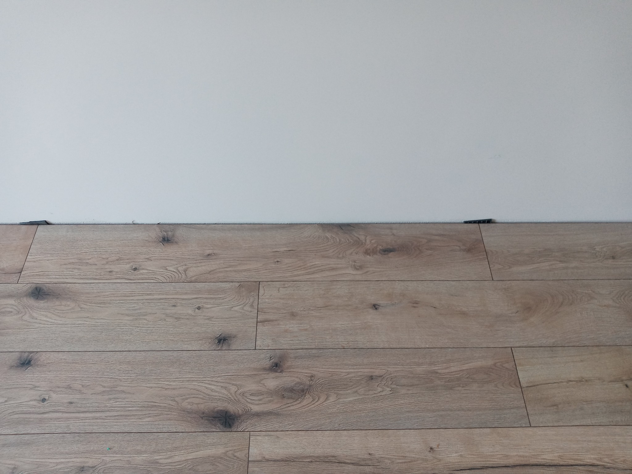 Fragment podłogi z paneli w odcieniach brązu, z widocznymi dystansami przy białej ścianie.