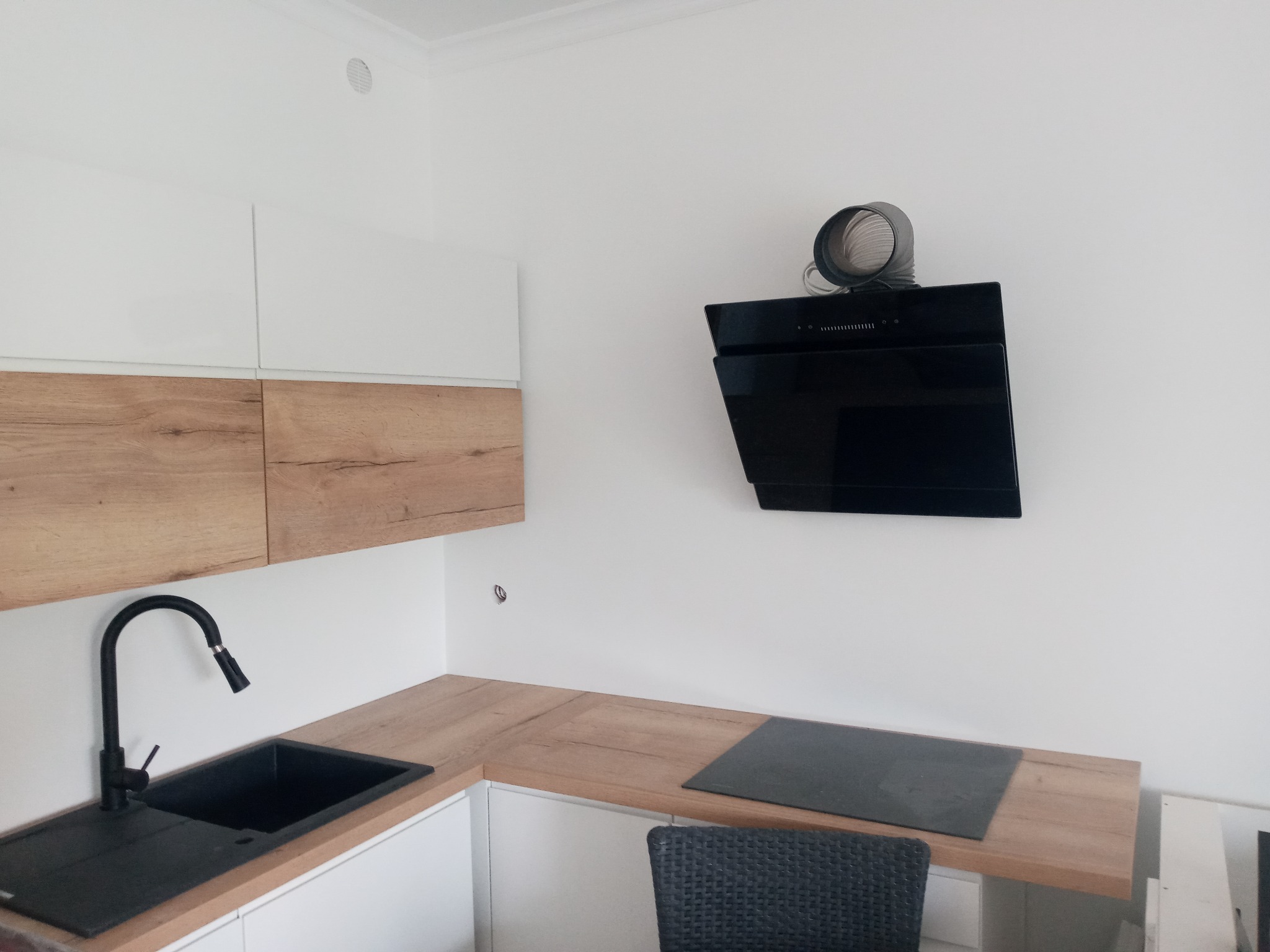 Nowoczesna kuchnia z białymi i drewnopodobnymi szafkami, czarnym zlewem i płytą indukcyjną pod czarnym okapem z widocznym przewodem wentylacyjnym.