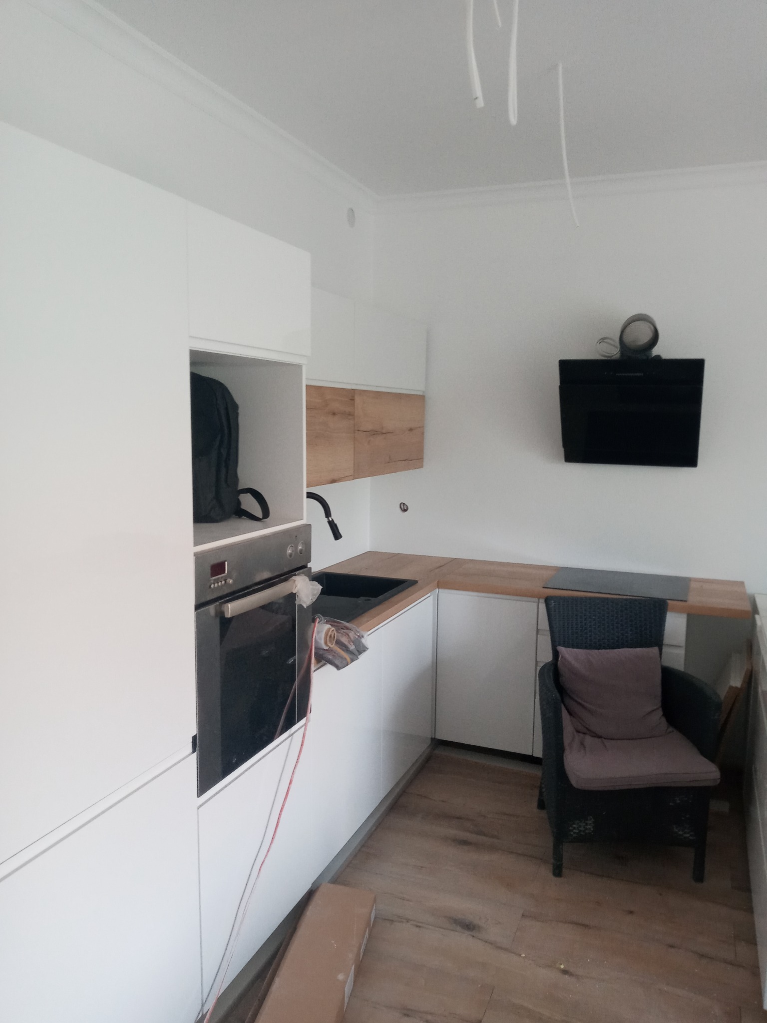 Nowoczesna kuchnia w trakcie montażu: białe szafki z drewnianymi akcentami, czarny okap, widoczne przewody elektryczne i karton na podłodze, czarne krzesło z poduszką w rogu.