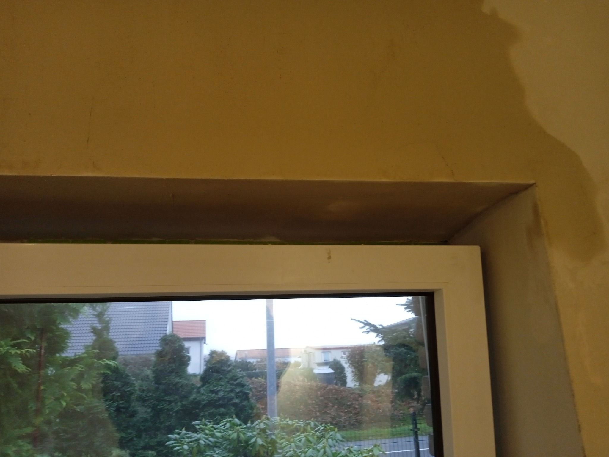 Surowa ściana wokół okna po szpachlowaniu, widoczne niedoskonałości i taśma malarska przy ramie okiennej, w tle zieleń i zabudowa mieszkalna.