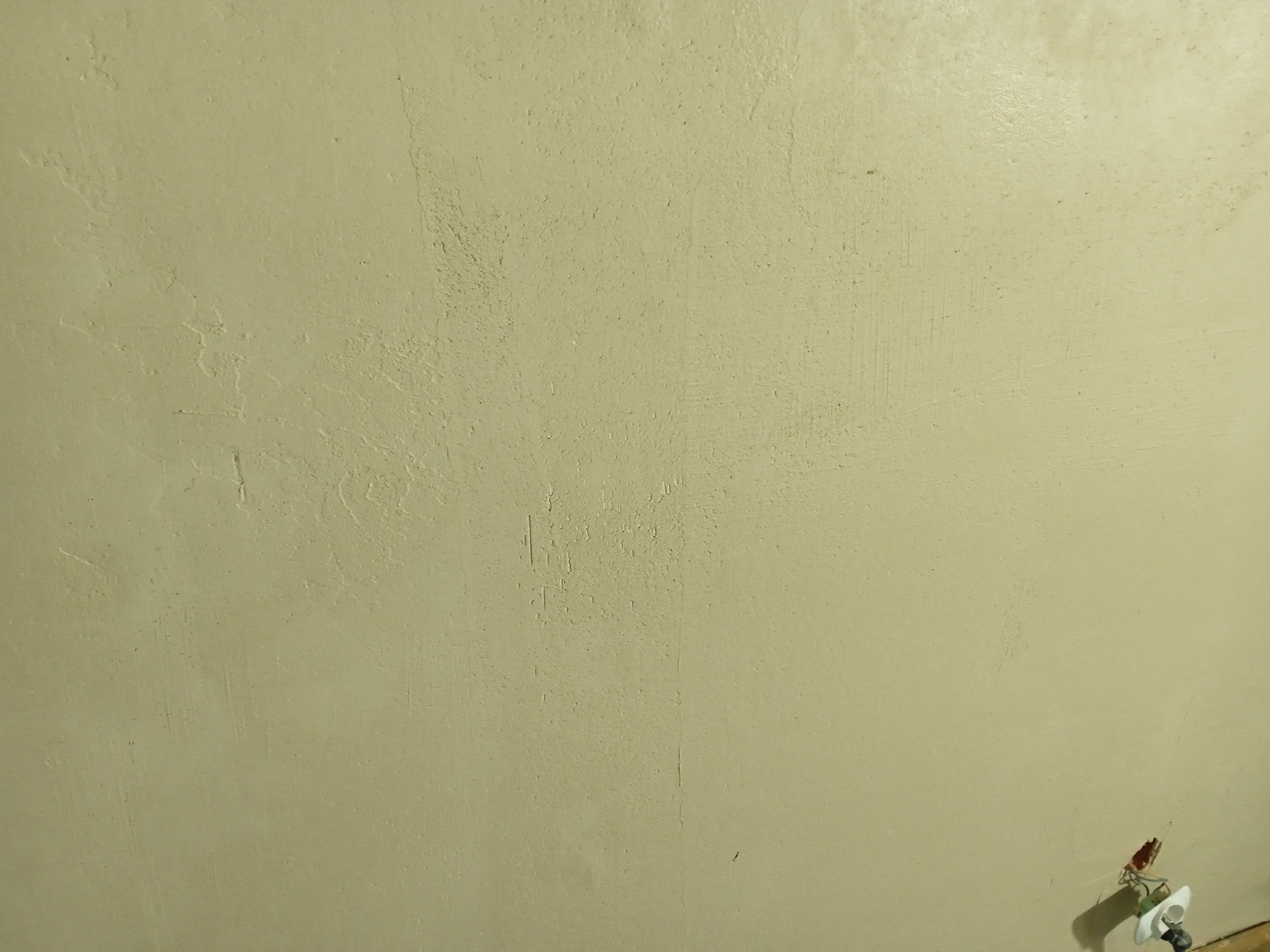 Ściana po szpachlowaniu, widoczne nierówności i faktura, przygotowana do malowania na jasny, kremowy kolor, z widoczną instalacją elektryczną w dolnym rogu.