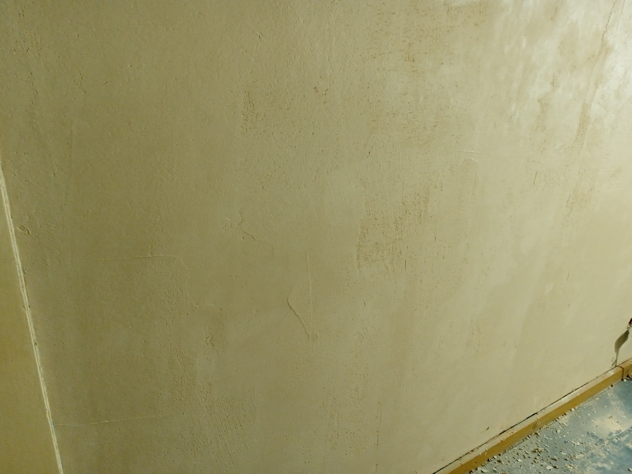 Ściana wewnętrzna po nałożeniu pierwszej warstwy szpachli w odcieniach beżu, widoczne niedoskonałości i nierówności faktury, listwa przypodłogowa z resztkami materiałów budowlanych na podłodze.