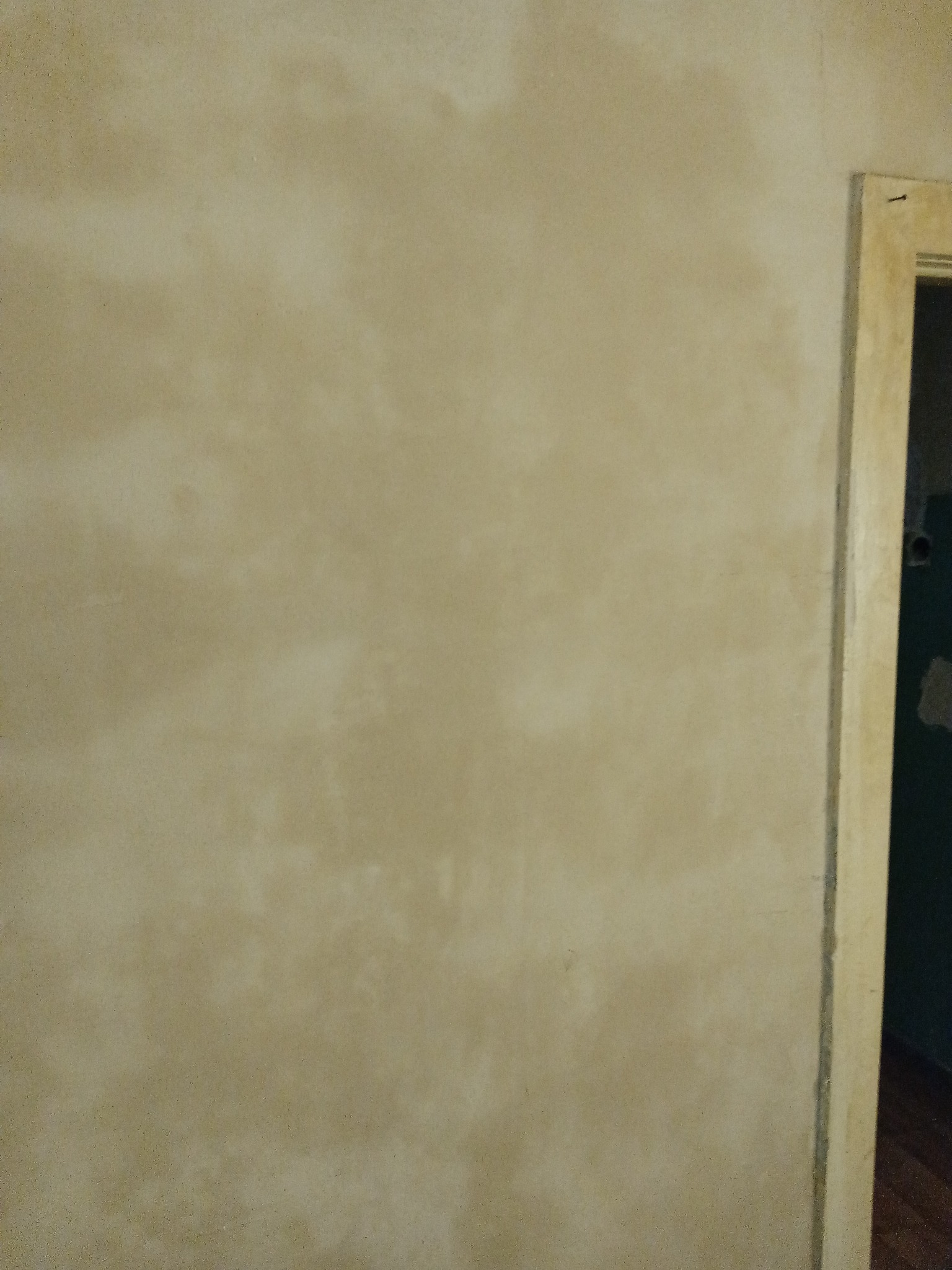 Ściana pokryta beżową, nierównomierną warstwą wykończeniową, widoczny fragment futryny drzwiowej po prawej stronie.