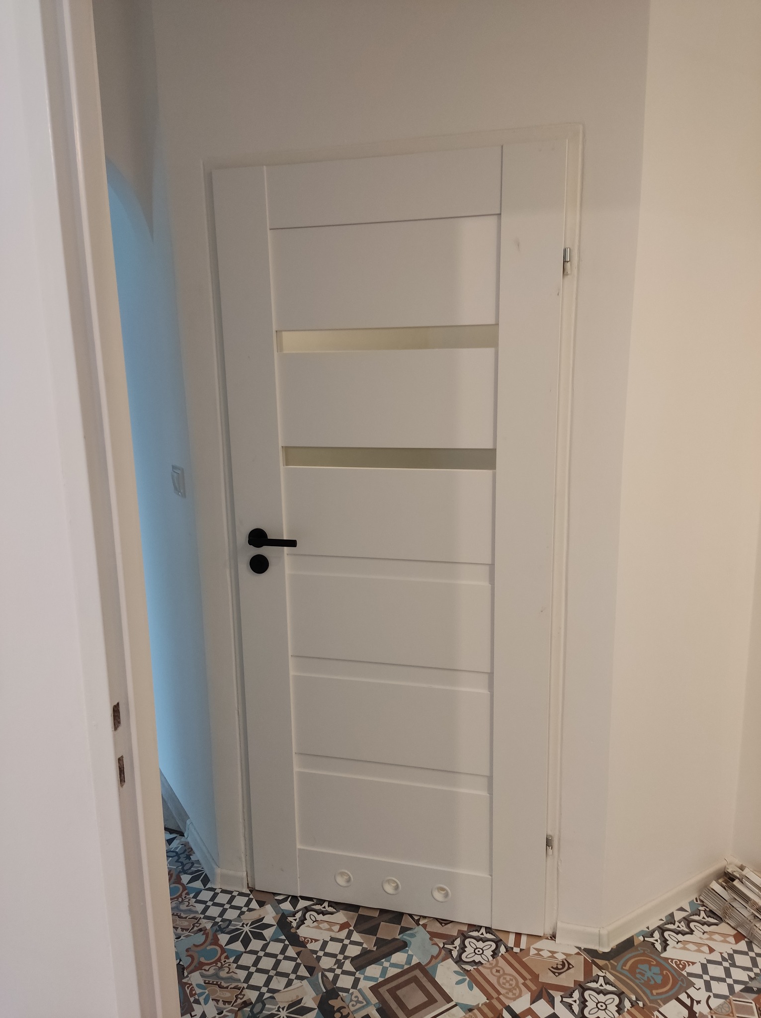 Białe drzwi wewnętrzne z poziomymi przeszkleniami i czarną klamką na tle wzorzystej podłogi w nowym mieszkaniu.