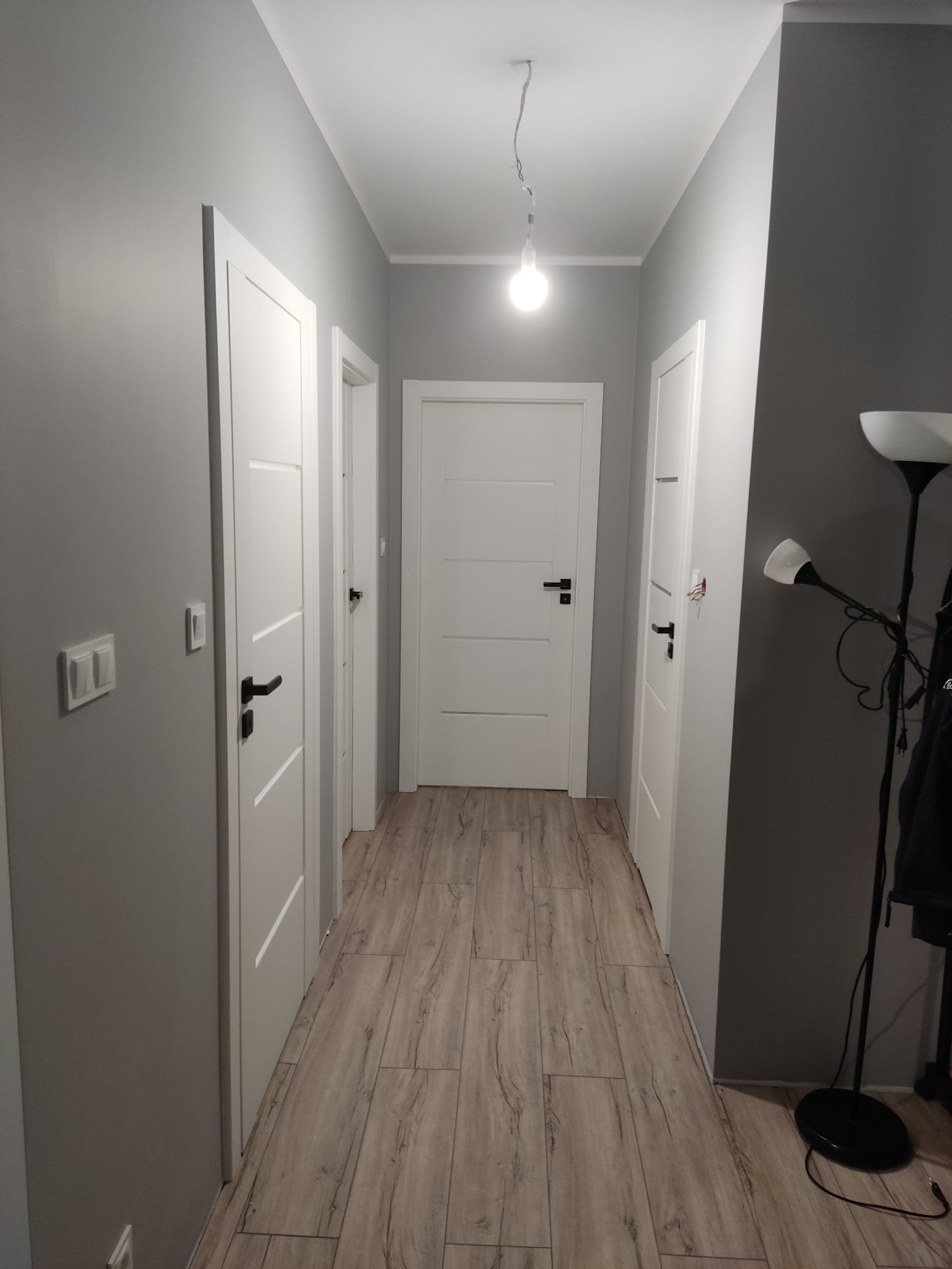 Korytarz z trzema białymi drzwiami z czarnymi klamkami, podłoga wyłożona panelami imitującymi drewno, szare ściany i wisząca pojedyncza żarówka.