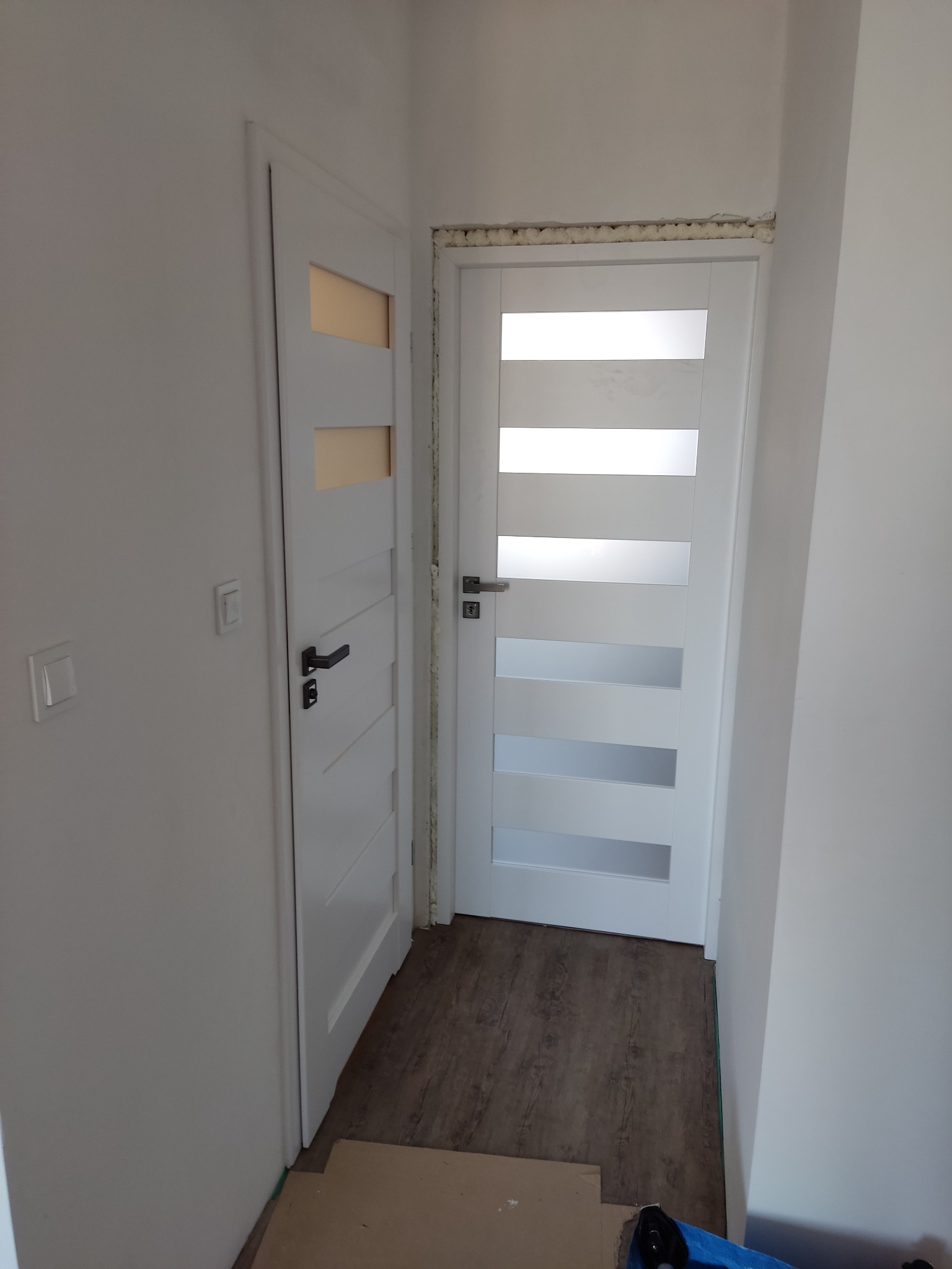 Dwa białe, nowoczesne skrzydła drzwiowe z przeszkleniami, zamontowane w białym korytarzu; widoczna pianka montażowa nad jednym z nich.