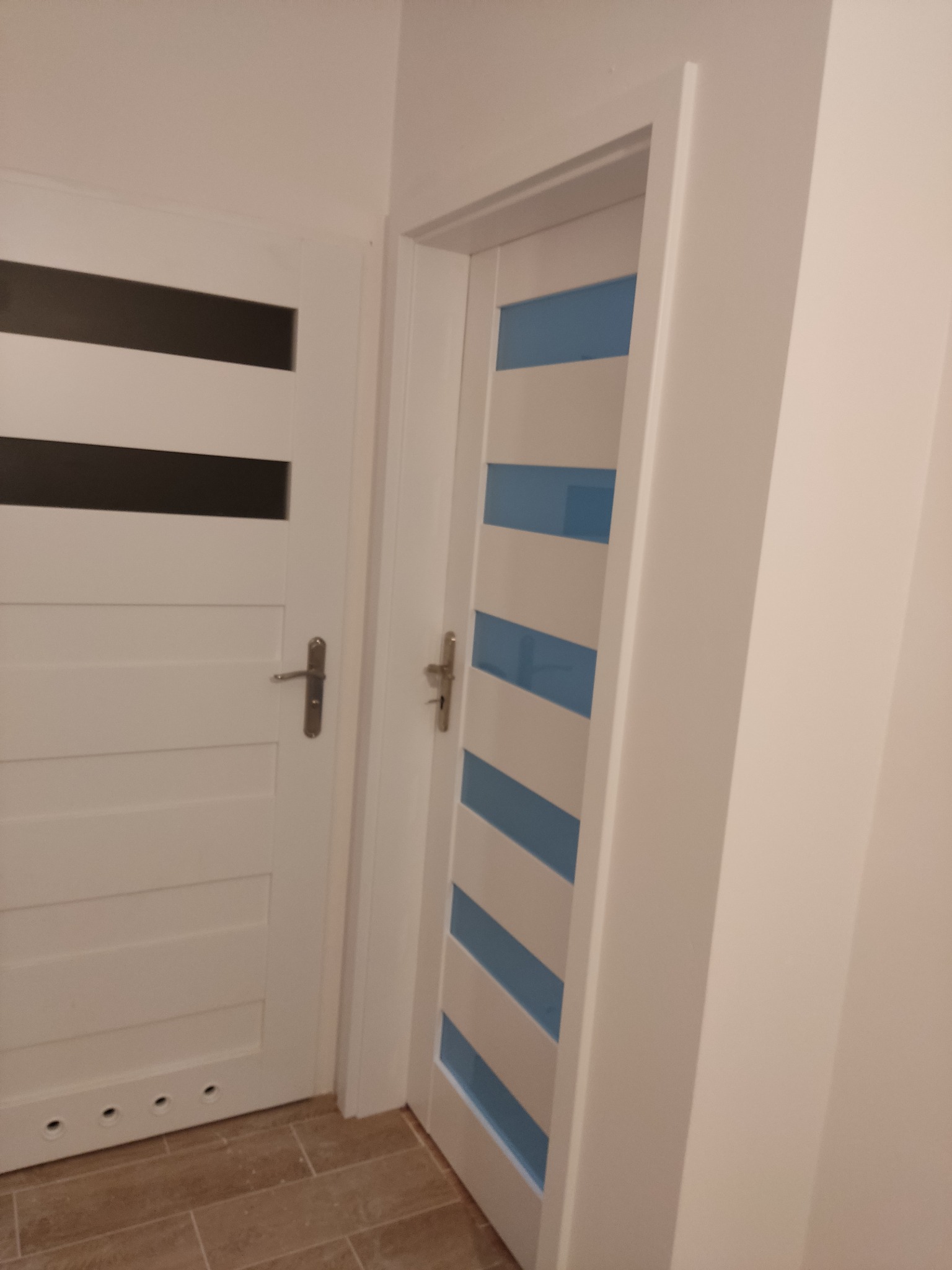 Dwa białe, wewnętrzne skrzydła drzwiowe z poziomymi przeszkleniami: jedno z ciemnymi szybami, drugie z błękitnymi, zamontowane w jasnym wnętrzu, widok z boku.