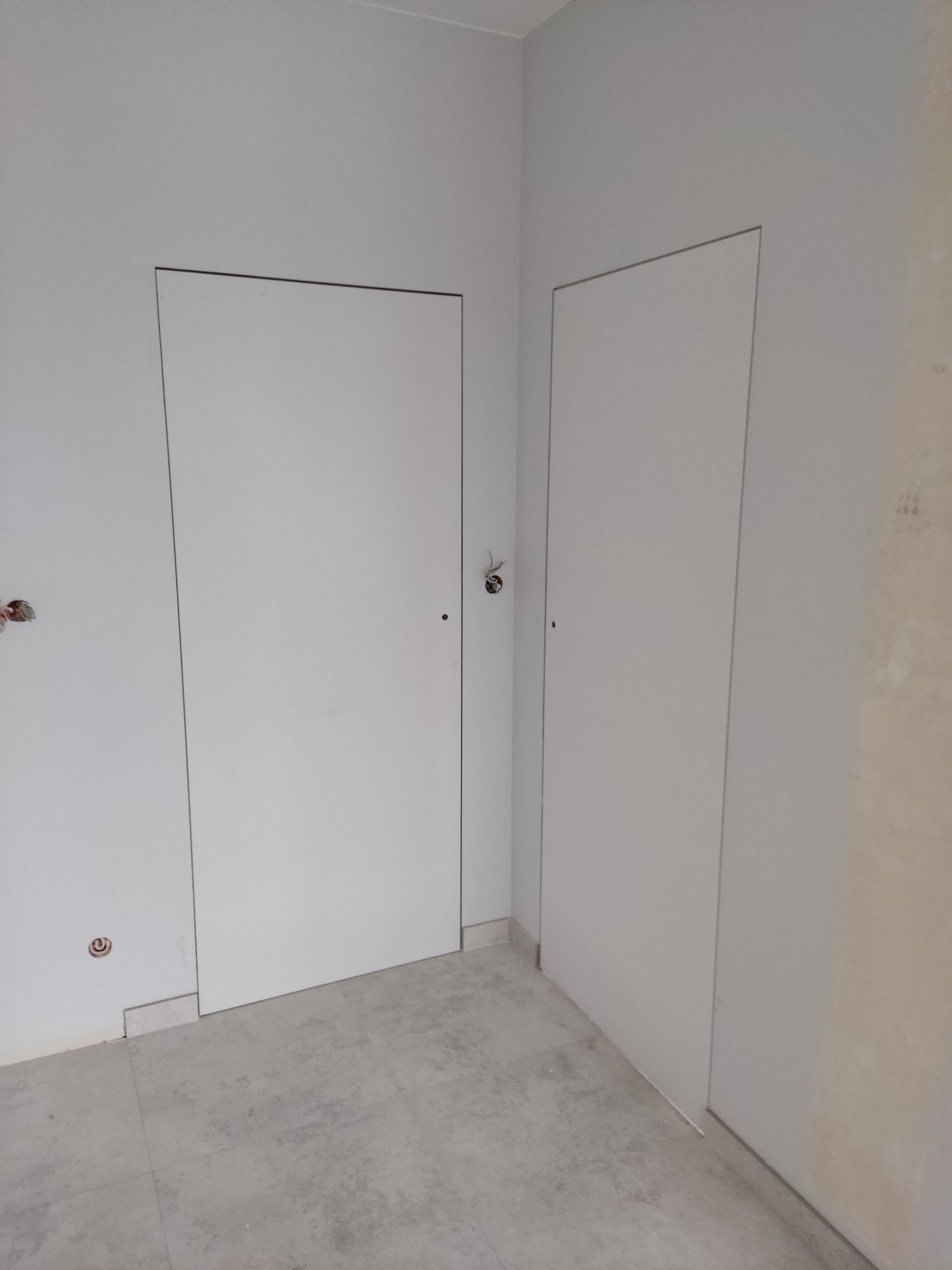 Dwie białe, ukryte drzwi zamontowane w szarej ścianie, widoczne gniazdka i surowe wykończenie podłogi.